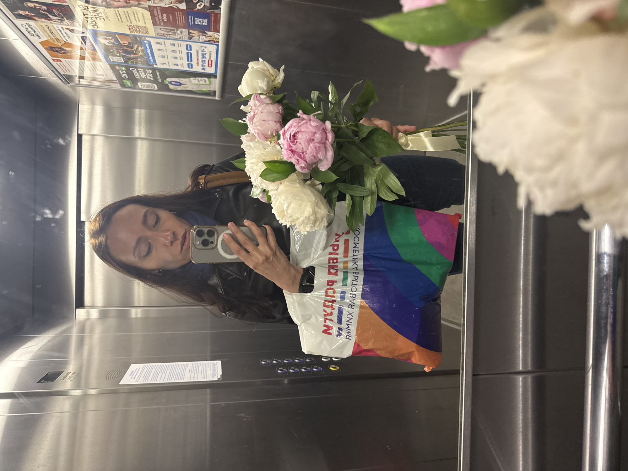 A woman is taking a selfie in an elevator with a bouquet of white and pink peonies, and a shopping bag. The text on the bag reads "RNMNX RABO". The background features the elevator's metal walls, elevator buttons, and informational posters. Based on the Cyrillic text visible in the image, the location is likely in the city of Saint Petersburg, Russia.