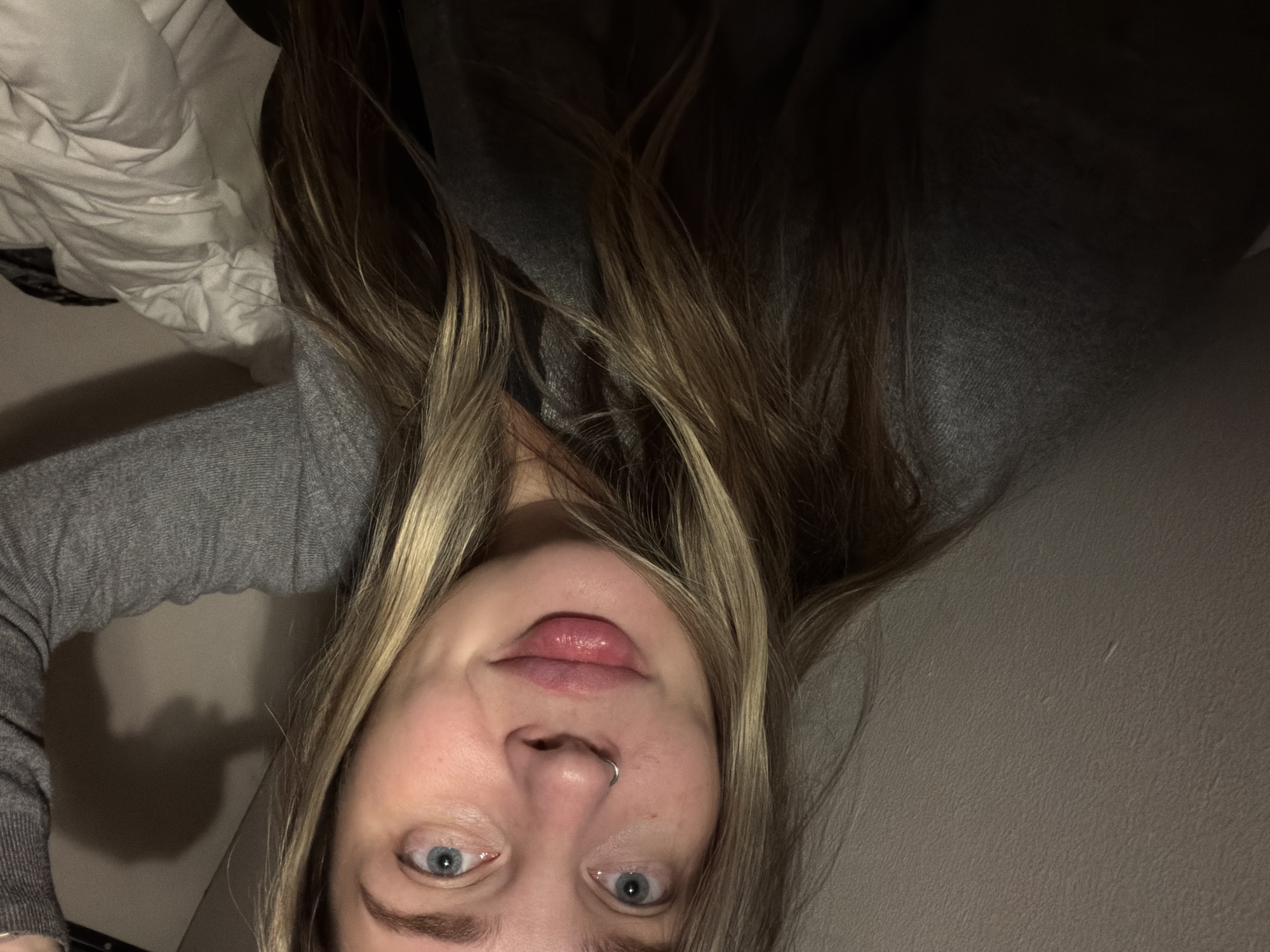 A self-portrait features a person, likely a woman, looking directly at the camera, appearing to recline indoors in Moscow, Russia. The image is oriented sideways. She has fair skin, wide-open light blue eyes, and long, multi-toned blonde and light brown hair framing her face. Her lips are slightly parted, revealing a glossy finish, and she has a small silver hoop piercing in her septum. She is wearing a grey knitted long-sleeved top.
The background is dimly lit, suggesting an evening or nighttime setting. To the left (top in the current orientation) is crumpled white fabric, possibly bedding or a blanket. To the right (bottom in the current orientation) is a plain, light-colored wall. Soft lighting illuminates her face from an unseen source, creating gentle shadows. Her expression is neutral and direct. No text is visible in the image.