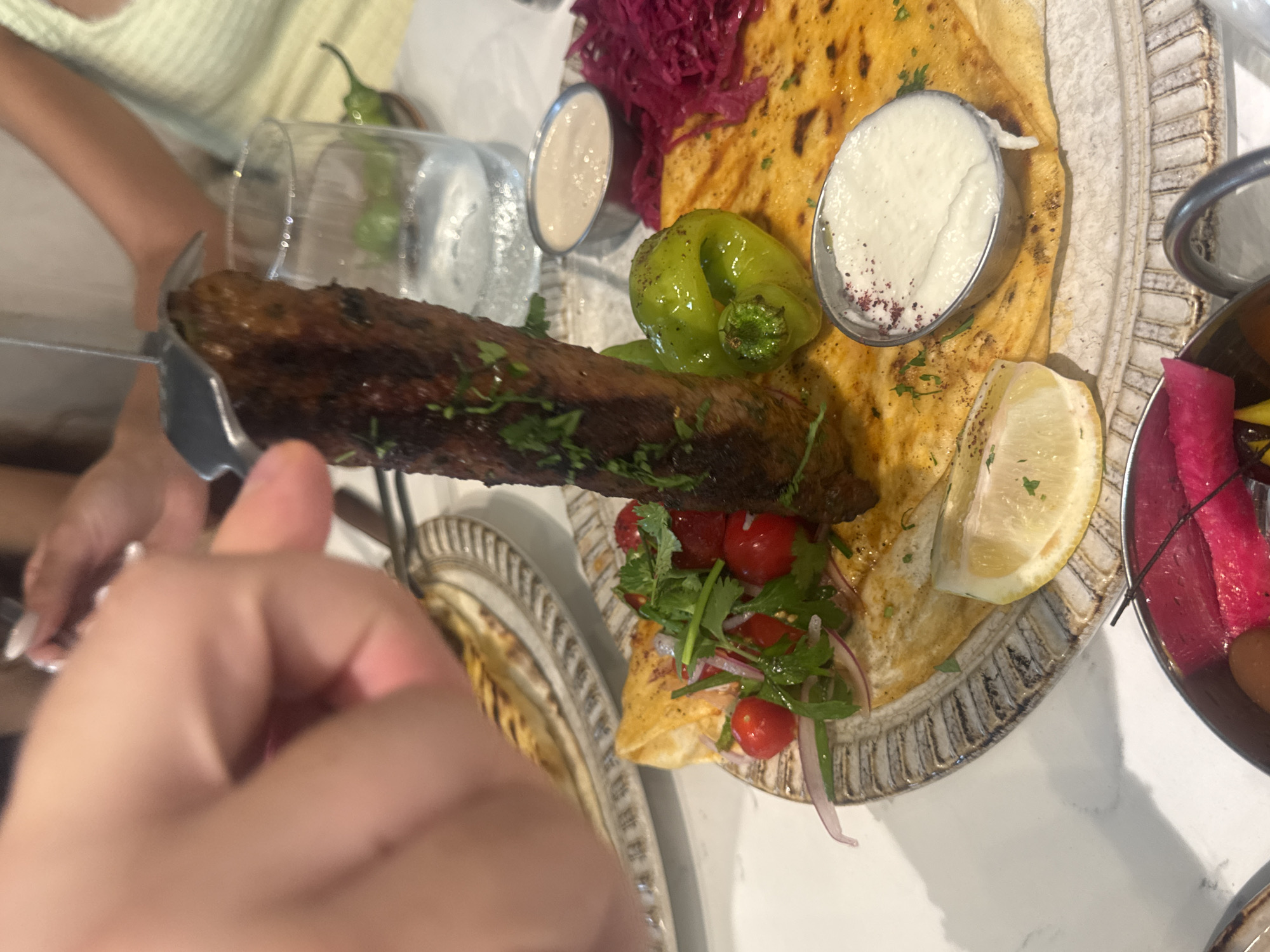 A close-up, high-angle shot shows a hand holding a fork that is spearing a grilled meat kebab. The kebab is brown and appears to be seasoned with herbs. It is served on a flatbread with a variety of accompaniments including a green bell pepper, cherry tomatoes, red onion, and leafy greens. There are also two small metal cups containing white dips, one sprinkled with red spice. A slice of lemon is visible on the side. The meal is presented on a decorative plate, which sits on a white, marble-like surface. In the background, a glass of water and a green chili pepper are visible, as are parts of other dishes and people's hands and arms. The scene suggests a restaurant setting, likely in Aventura, Florida, USA.