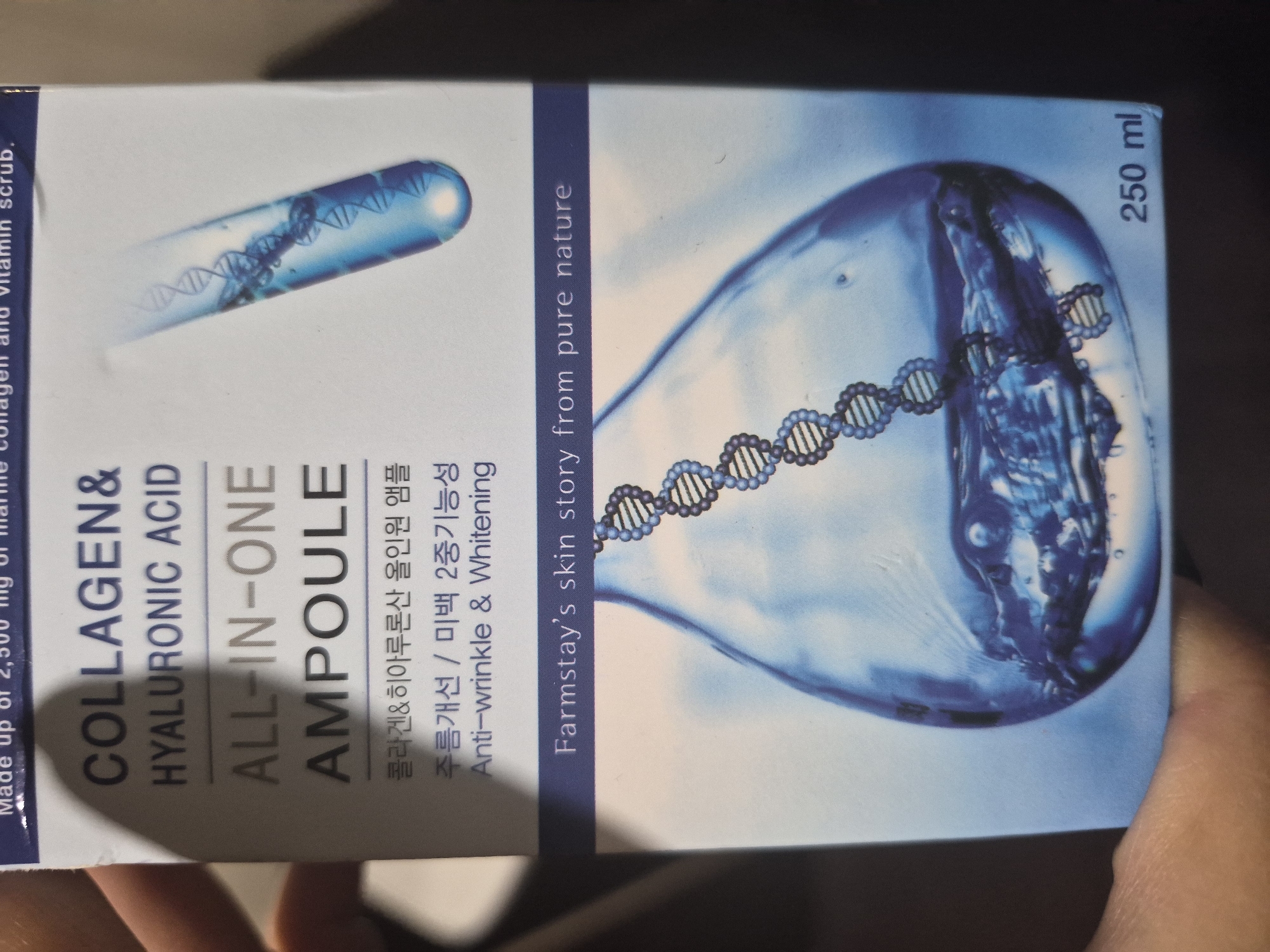 A hand holds up a blue and white product box for a skincare item, likely an ampoule or serum. The box is oriented vertically, and the image is taken indoors with a dark, out-of-focus background.
The front of the box prominently displays the product name "COLLAGEN & HYALURONIC ACID ALL-IN-ONE AMPOULE" in large, dark letters. Below this, the Korean translation "콜라겐&히아루론산 올인원 앰플" is visible. Further down, "Anti-wrinkle & Whitening" is stated, alongside its Korean counterpart "주름개선 / 미백 2중기능성" (Wrinkle Improvement / Whitening 2-in-1 function).
The design features scientific-looking graphics: a test tube or ampoule shape containing a blue liquid and DNA strands on the left, and a larger, tear-drop shaped liquid mass with DNA strands on the right. A blue vertical strip running down the middle states, "Farmstay's skin story from pure nature." The volume of the product, "250 ml," is printed vertically on the right side. Partially visible text on the far left edge of the box suggests the product contains "2,300 mg of marine collagen and vitamin scrub."
The hand holding the box is light-skinned, with visible fingers on the right side of the frame, indicating the user is presenting the product. The lighting is artificial, illuminating the box clearly against the dim background. The scene gives no specific clues about the time of day or location within Yerevan, Armenia, other than it being indoors.