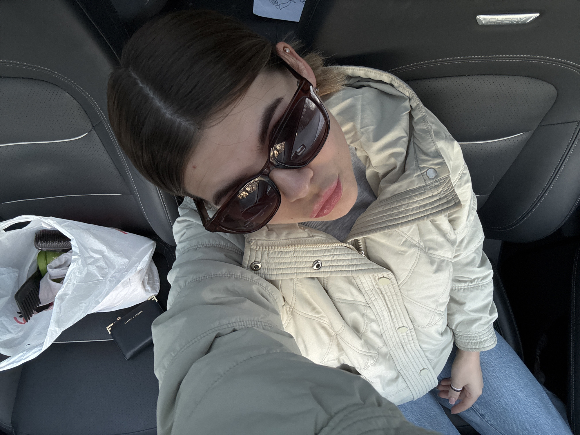 An overhead high-angle selfie depicts a young adult female seated in the passenger seat of a vehicle. The subject has dark brown hair parted centrally and wears dark brown sunglasses, a light beige quilted jacket with a zippered front and snap buttons, a grey t-shirt, and blue jeans. Her left hand, adorned with a silver-colored ring, rests on her upper leg. To her left, on the black leather car seat, rests a crumpled white plastic shopping bag containing a black wide-tooth comb, a black hairbrush, and a green cylindrical container. Beneath the bag, a black wallet with "CLASSIC & MODERN" text and a gold-colored clasp is visible. The car seat features white stitching and perforated sections, with a chrome "CHERY" emblem affixed to the right side of the seatback. The precise location within the city of Chaikovsky, Russia, cannot be determined from the visual evidence provided in the image.