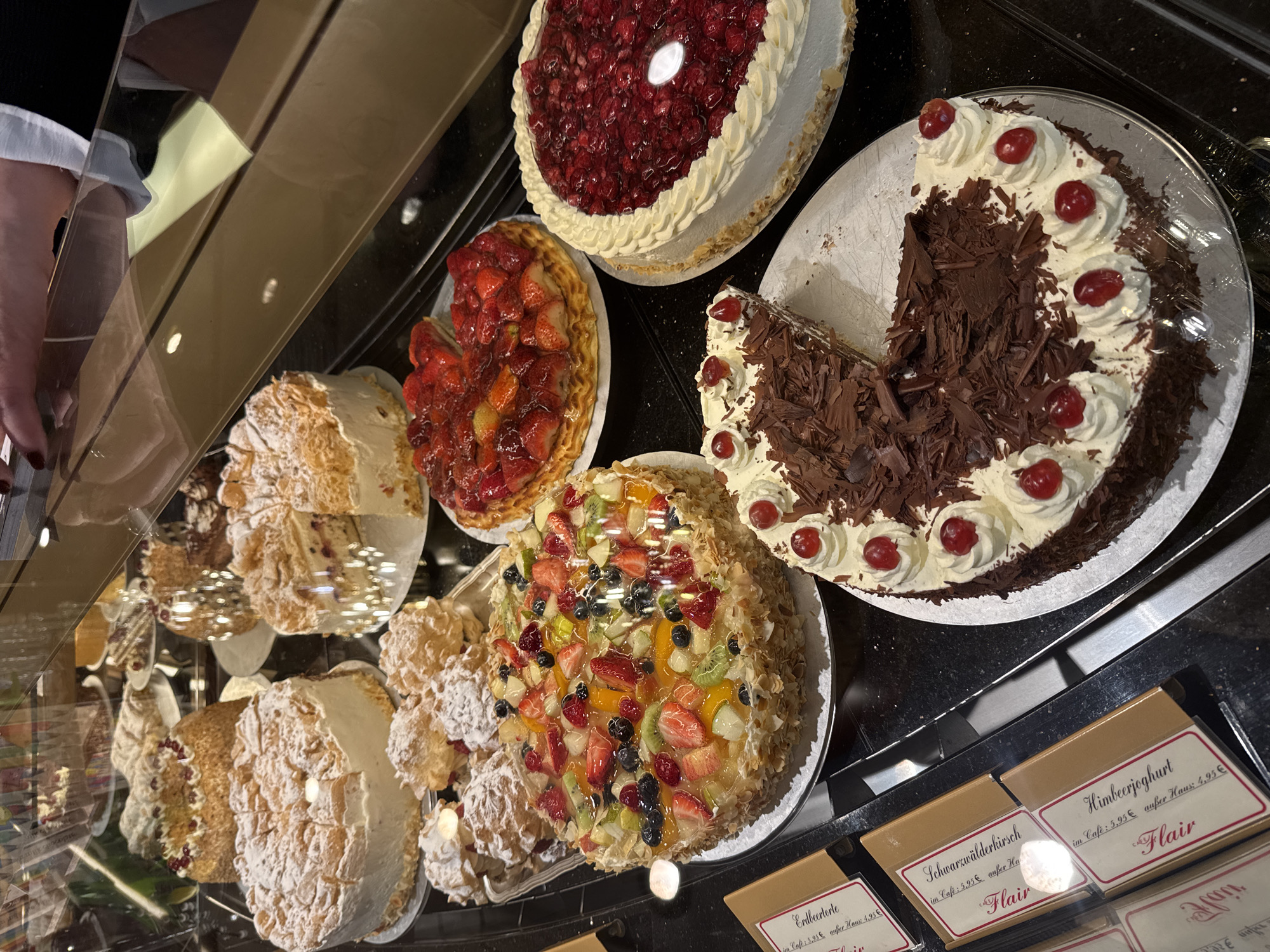 The image captures a display case filled with various cakes and pastries, likely within a bakery or café in Cochem, Germany. The cakes are arranged on plates and include Black Forest cake (Schwarzwälderkirsch), strawberry cake (Erdbeertorte), raspberry yogurt cake (Himbeerjoghurt), and other fruit-topped cakes. Price tags are visible below the cakes, indicating that the cakes are for sale. A portion of a person's arm, possibly a store clerk, is visible on the left side of the image. The reflective glass of the display case is visible.