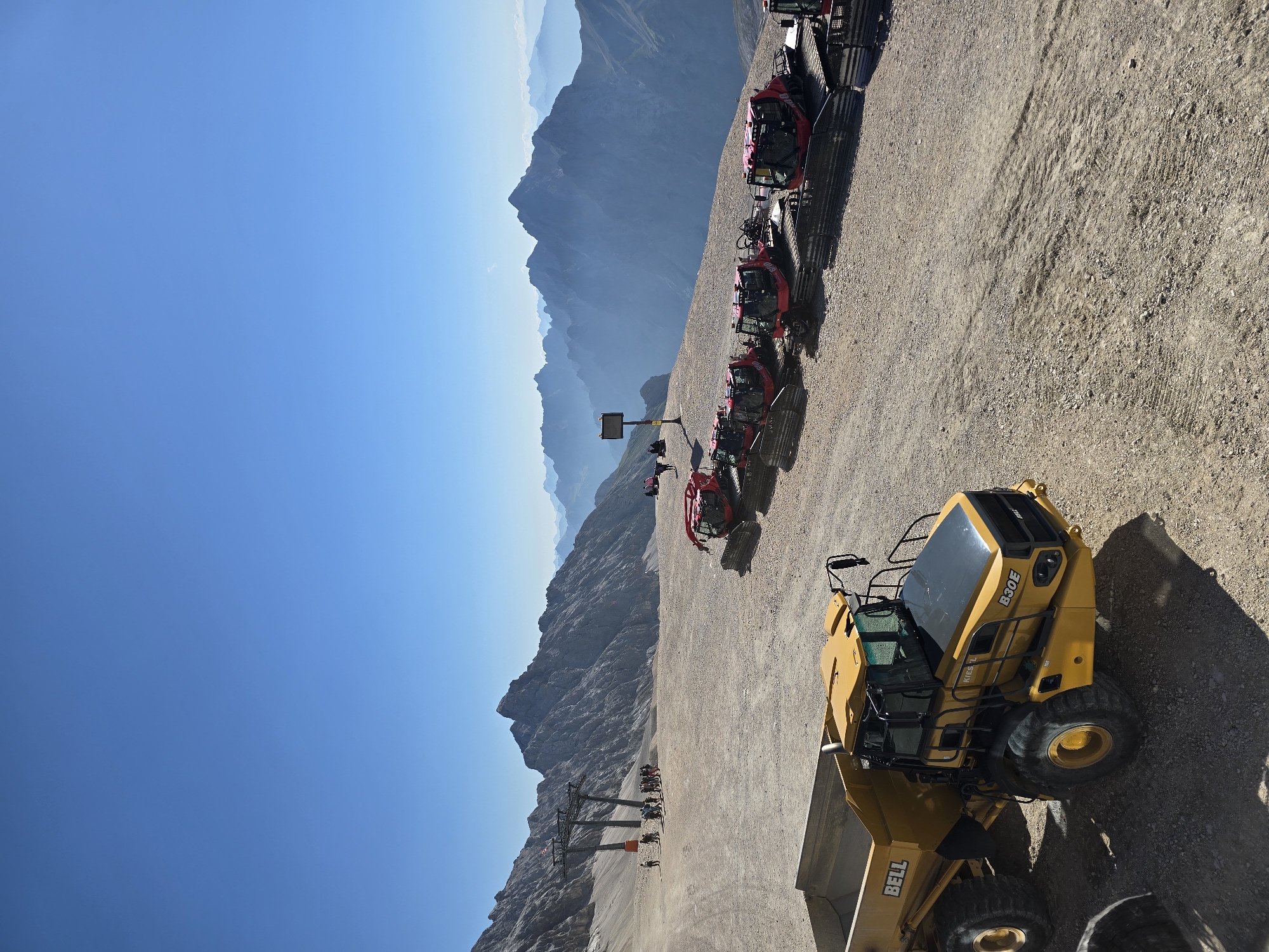 This image captures a scenic mountain landscape with several vehicles and people present. The scene is set on a high-altitude terrain, likely a plateau or a mountain pass, given the vast expanse of rocky ground and the distant, hazy mountain ranges. The sky is a clear, bright blue, indicating a sunny day.
In the foreground, a bright yellow articulated dump truck, identified by the "BELL" logo on its side and "B30E KIESEL" on the front, is parked on a dusty, gravelly surface. Its large wheels and heavy-duty construction suggest it's used for construction or heavy transport in rugged environments.
To the right of the dump truck, a line of red, tracked vehicles, possibly snow groomers or maintenance equipment, are parked. These vehicles have a distinctive front plow or tiller and are designed for working on snow or rough terrain.
In the mid-ground, a group of people are gathered near a ski lift structure, which consists of large metal pylons and cables disappearing up the mountainside. The people appear to be tourists or workers, some standing and observing, while others are engaged in conversation.
The background is dominated by imposing mountain peaks, layered and fading into a soft blue haze, indicating significant distance. The overall environment is mountainous and appears to be an outdoor recreation or construction site at a high elevation. The location is likely the Zugspitze area near Garmisch-Partenkirchen, Germany, given the presence of ski lift infrastructure and the characteristic Alpine scenery.