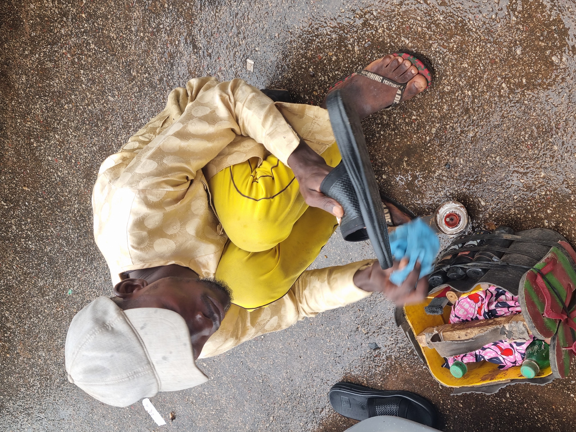 A man lies on a wet paved surface in Jos, Nigeria. He wears a white baseball cap, a light yellow patterned long-sleeved shirt, and yellow pants. He has a pair of sandals on his right foot. In his hands, he holds a black sandal and a light blue cloth. A yellow container filled with assorted items, including fabric, bottles, and more sandals, sits beside him.