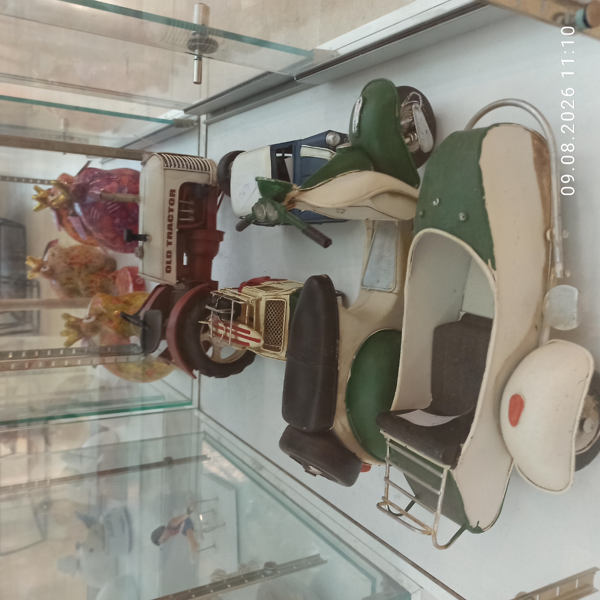 In the city of Molfetta, Italy, a collection of vintage toy vehicles is displayed inside a glass cabinet. The toys include a red and white tractor with "OLD TRACTOR" printed on its side, a white and green scooter with a sidecar, and a blue and white three-wheeled vehicle that appears to be a tuk-tuk. In the background, other decorative objects, possibly ceramic figurines, are visible, some adorned with crown-like embellishments. The scene is illuminated by artificial light, casting subtle reflections on the glass surfaces of the display case. The floor appears to be a light-colored surface.
