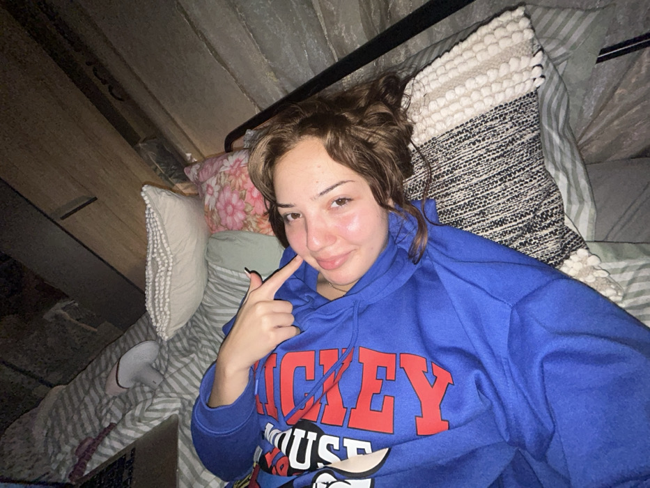 A woman lies on a bed with several pillows and striped sheets. She is wearing a blue Mickey Mouse sweatshirt and points to her cheek with a black fingernail. Her brown hair is styled loosely. The space above and to the side appears to be an attic room or sloped ceiling. One white slipper is visible on the bed.