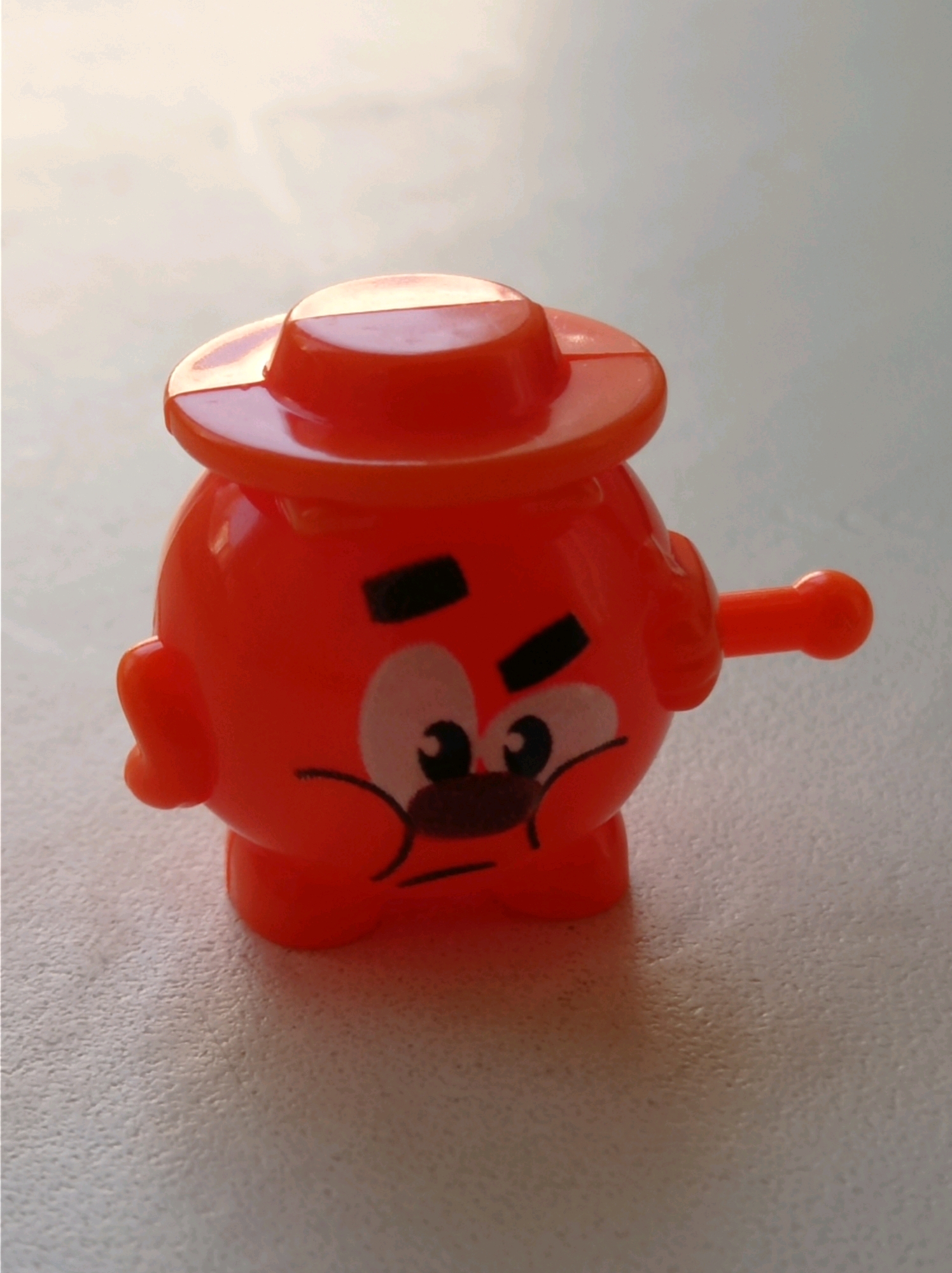 The image shows a close-up, eye-level shot of a small, bright orange plastic toy. The toy is spherical with a flat-topped hat on its head. It has two black, oval-shaped eyes with white pupils and black irises. Below the eyes are two black, curved lines representing cheeks or a puffed-out mouth. A small, orange projection extends from the right side of the toy's body. The toy is positioned on a textured, light-colored surface that appears to be a countertop or table. The background is softly blurred and lighter in tone. There is no visible indication of the city of Degtyarsk, Russia, or any specific location.