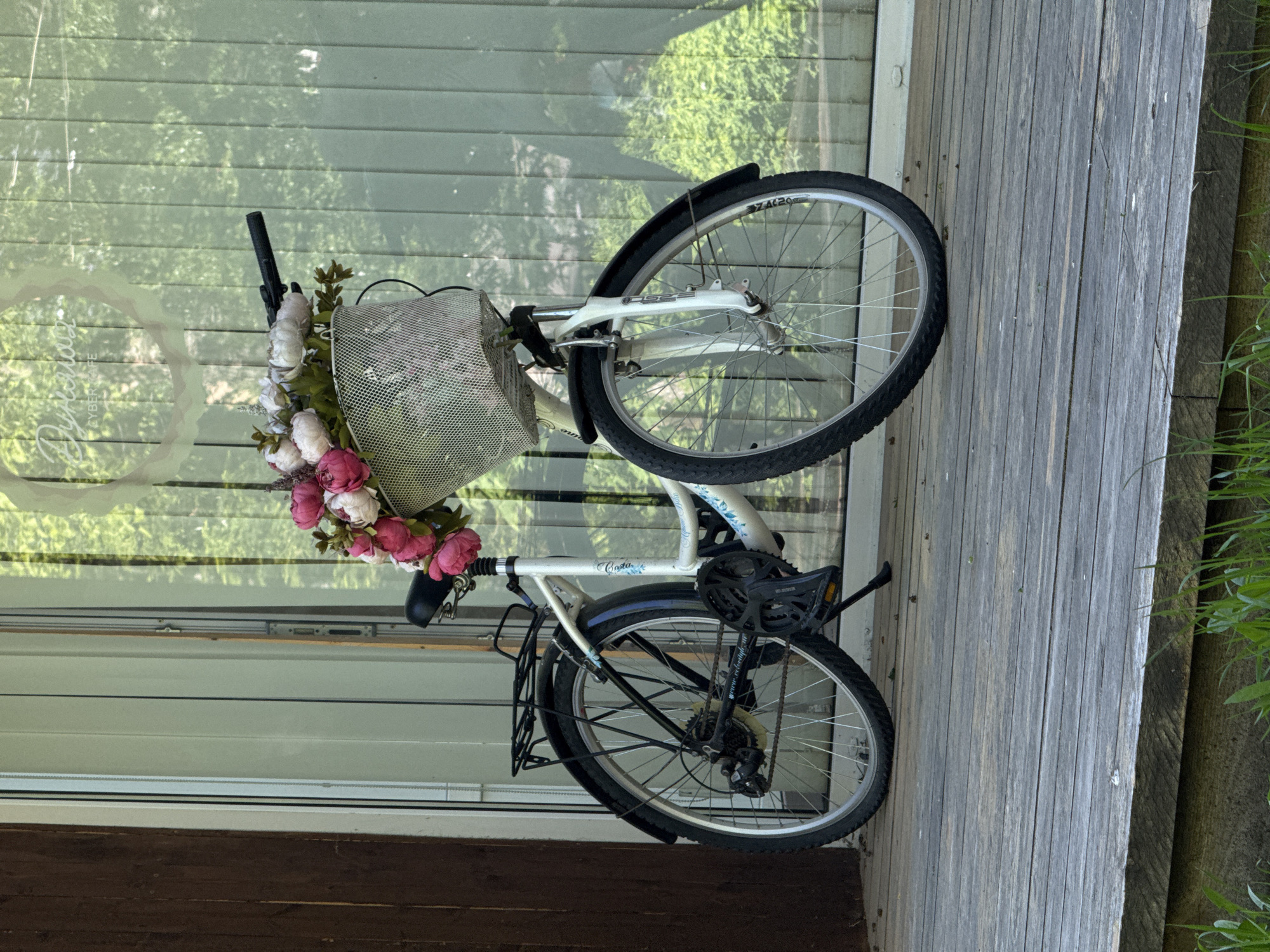 A white bicycle with a basket filled with pink and white flowers is leaned against a glass-paneled wall and a wooden plank surface. The location is Perm, Russia, indicated by the Cyrillic text "Сделано в Перми" (Made in Perm) visible on a circular sticker on the glass. The bicycle is equipped with front and rear fenders, and a rear rack. The background shows a blurred outdoor scene with greenery.