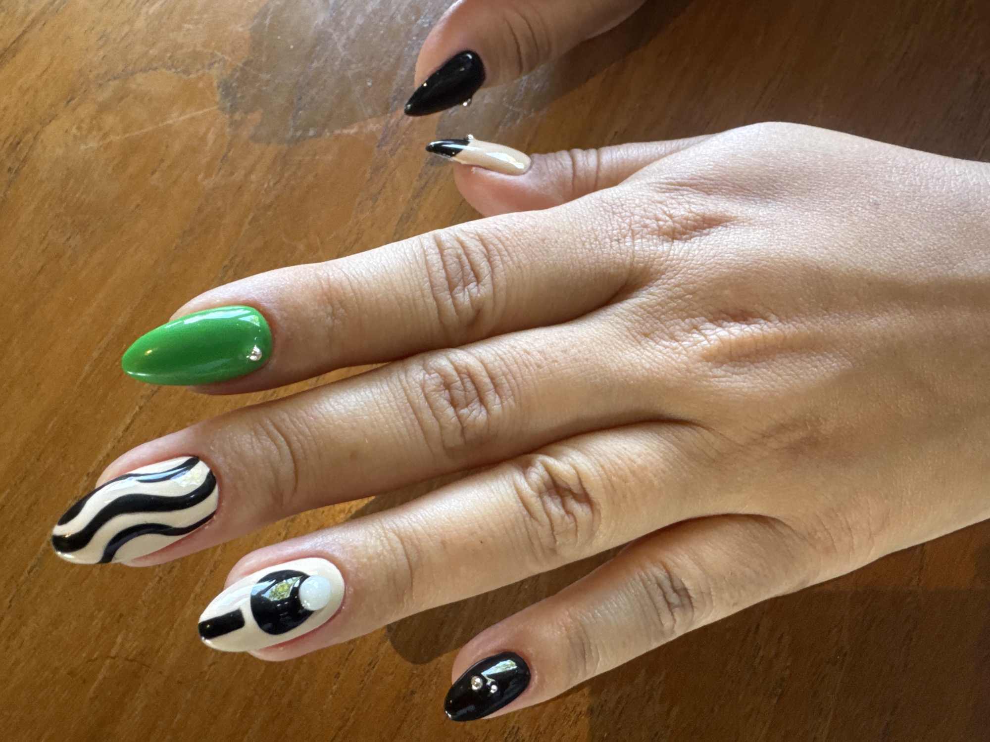 A photograph depicts a right hand with painted nails, resting on a wooden surface. The nails are painted in different colors and designs: black, green, beige, white, and a zebra-striped design. Some nails have small rhinestones. The location is inferred to be North Kuta, Indonesia. No additional subjects, actions, or interactions are visible. The environment consists only of the hand and wooden surface.