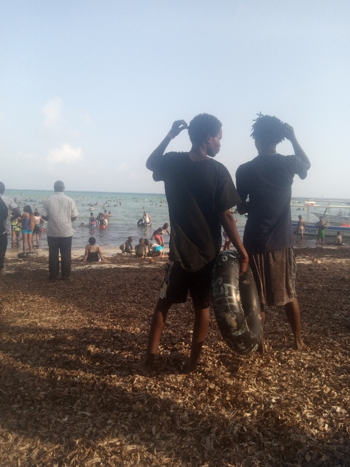This image depicts a crowded coastal scene in Shanzu Ward, Kenya, under a clear, light blue sky with minimal cloud cover.
The foreground consists of a beach covered extensively with a layer of dried, brown organic matter, likely seaweed or plant debris. Visible shadows suggest sunlight originating from the right side of the frame. Two young males stand in the immediate foreground, observed from behind, facing the ocean. The male on the left wears a dark t-shirt and dark shorts, with his right hand raised to his head, appearing to adjust his hair. The male on the right wears a dark t-shirt and brown shorts, also with his left hand raised to his head, adjusting his hair. He holds a dark, possibly deflated or partially deflated inflatable ring with visible white markings against his left leg.
In the midground, a diverse group of individuals, including adults and children, occupies the shoreline and the shallow, light blue-green water. One adult male, seen from behind, stands prominently in a light plaid shirt and dark trousers. Several people are wading, sitting, or playing in the water near the shore. Many individuals in the water are utilizing inflatable rings.
Further into the background, a large number of people are visible submerged in the water. Several small watercraft, likely boats, are present near the horizon line on the right side of the image. The overall environment suggests a recreational beach setting with numerous people engaged in water activities.