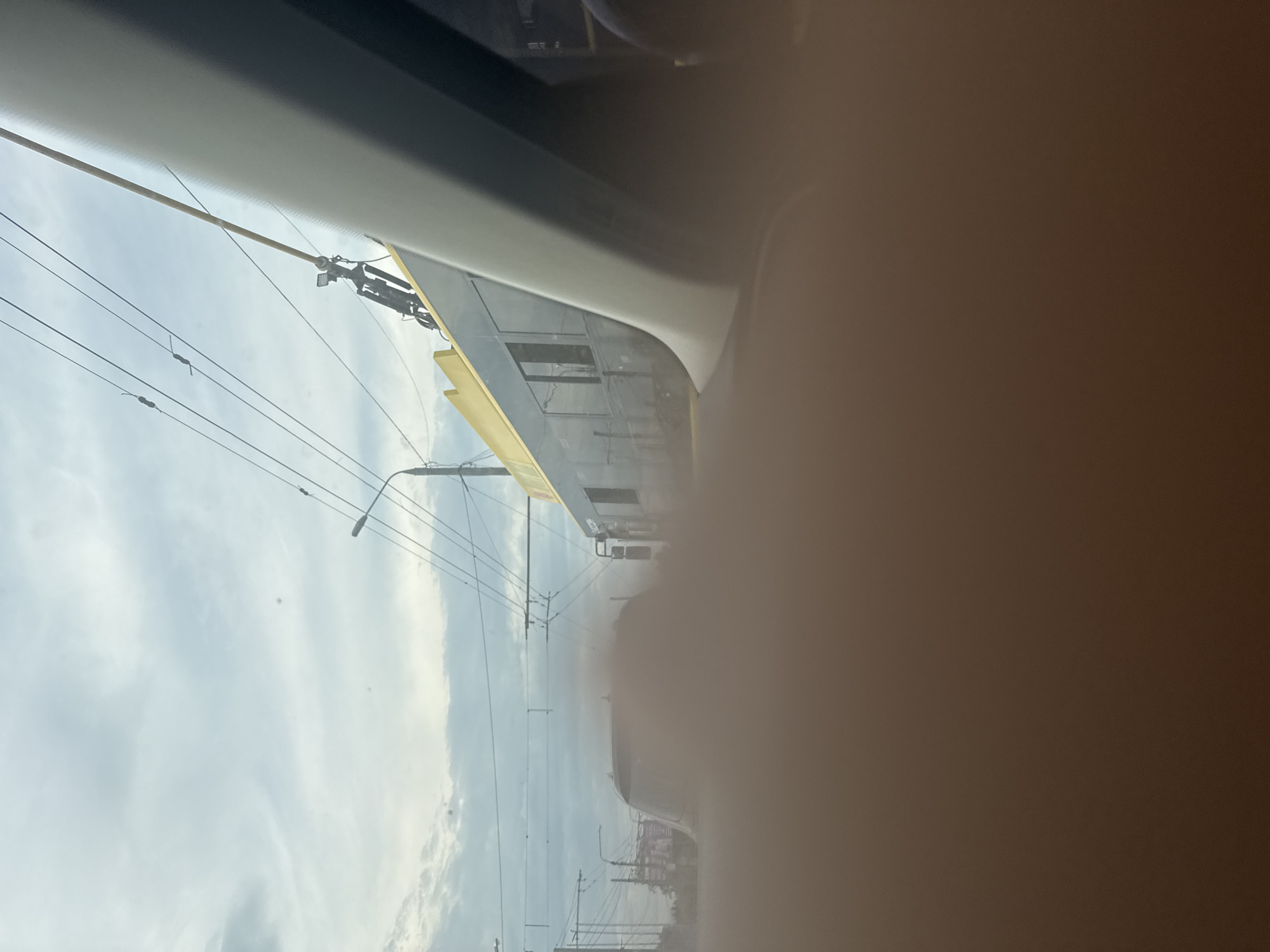 The image presents an urban street scene captured from the interior of a vehicle, looking out the right-side window. A significant portion of the lower frame is obscured by a blurry, brown-beige surface, likely the vehicle's dashboard or door panel. A light grey vehicle pillar extends diagonally from the top right into the mid-right, partially framing the view.
In the midground, a dark-colored trolleybus with a distinct yellow stripe along its roofline is depicted in motion, traveling parallel to the observer's vehicle. Its trolley pole extends upwards, connecting to a complex network of overhead electrical lines that traverse the sky. Multiple dark-tinted windows are visible along the side of the trolleybus. Beyond the trolleybus, a blurry, dark-colored car is partially visible in the traffic flow.
The background features an overcast sky characterized by light grey and white cloud formations. Numerous utility poles and intersecting power lines are prominent across the upper section of the image, alongside a visible streetlight. The scene conveys a bustling urban environment with public transportation infrastructure, with no discernible individuals or specific city identifiers.