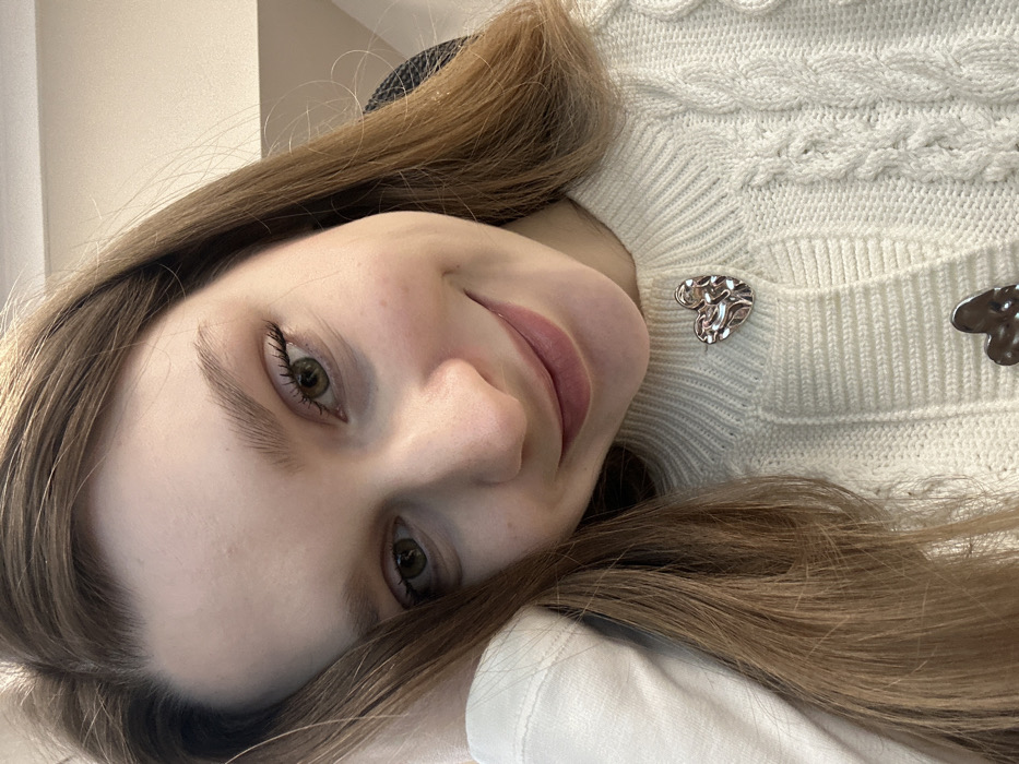 A young woman with light skin and long, light brown hair smiles directly at the camera. Her eyes are a greenish-hazel. She is wearing a cable-knit white sweater adorned with two silver heart-shaped buttons. The photo is taken from an unusual angle, tilted so that her face is diagonal within the frame. A hint of a dark chair or headrest is visible in the background, suggesting she might be seated. Her expression is warm and engaging.