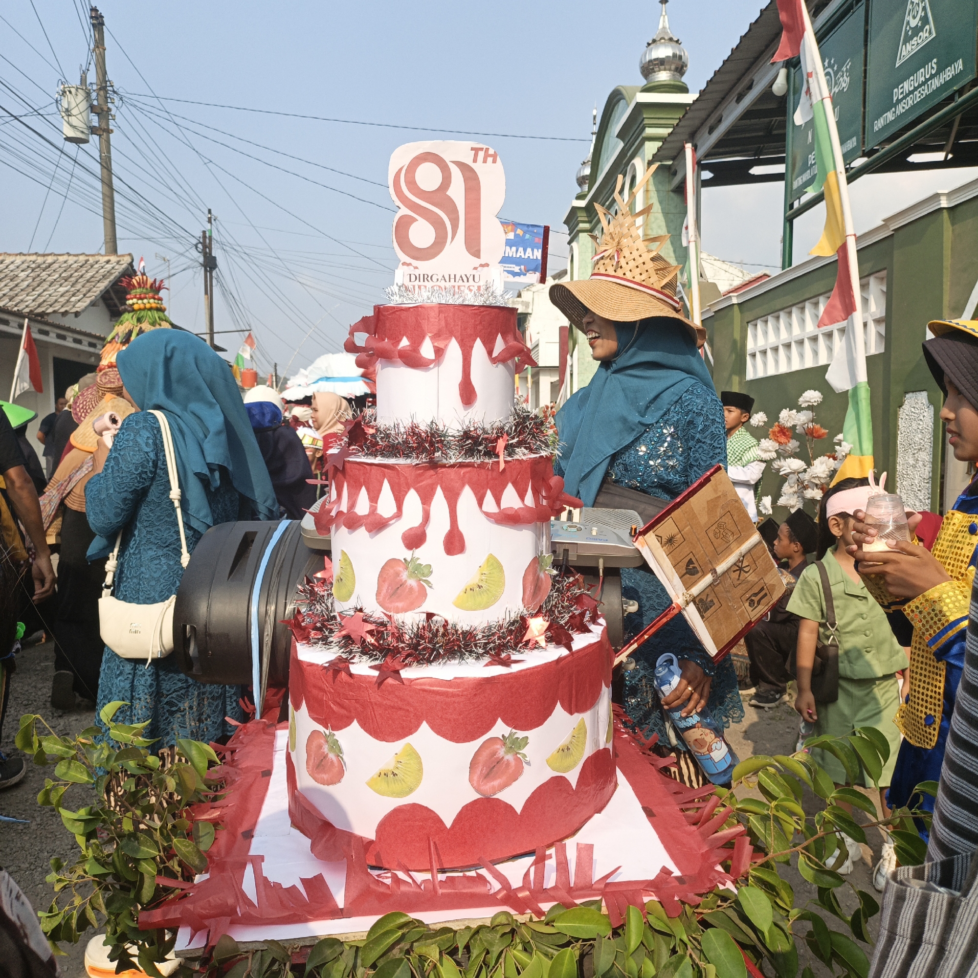 The image depicts a street scene in Tanahbaya, Indonesia, possibly during a celebratory event or parade. A large, tiered cake-like structure, decorated with red paper frills and images of strawberries and kiwi slices, is a central focus. The cake is adorned with a large sign displaying "81" and the text "DIRGAHAYU INDONESIA," suggesting a celebration of Indonesia's Independence Day or a similar national anniversary.
Several people are visible, most of them women wearing headscarves (hijabs). One woman, wearing a blue hijab and a decorated straw hat, is standing next to the cake, smiling and holding a small book-like object made of cardboard. Another woman in a blue lace dress and a light-colored purse is seen holding a blue water bottle with a cartoon character on it. Children are also present in the background, some wearing traditional attire.
The environment suggests an outdoor setting on a sunny day. Buildings with green facades and decorative elements are visible in the background, along with utility poles and wires. Indonesian flags, in red and white, are displayed on poles and buildings. The ground appears to be a paved road. The overall atmosphere seems festive and communal.
