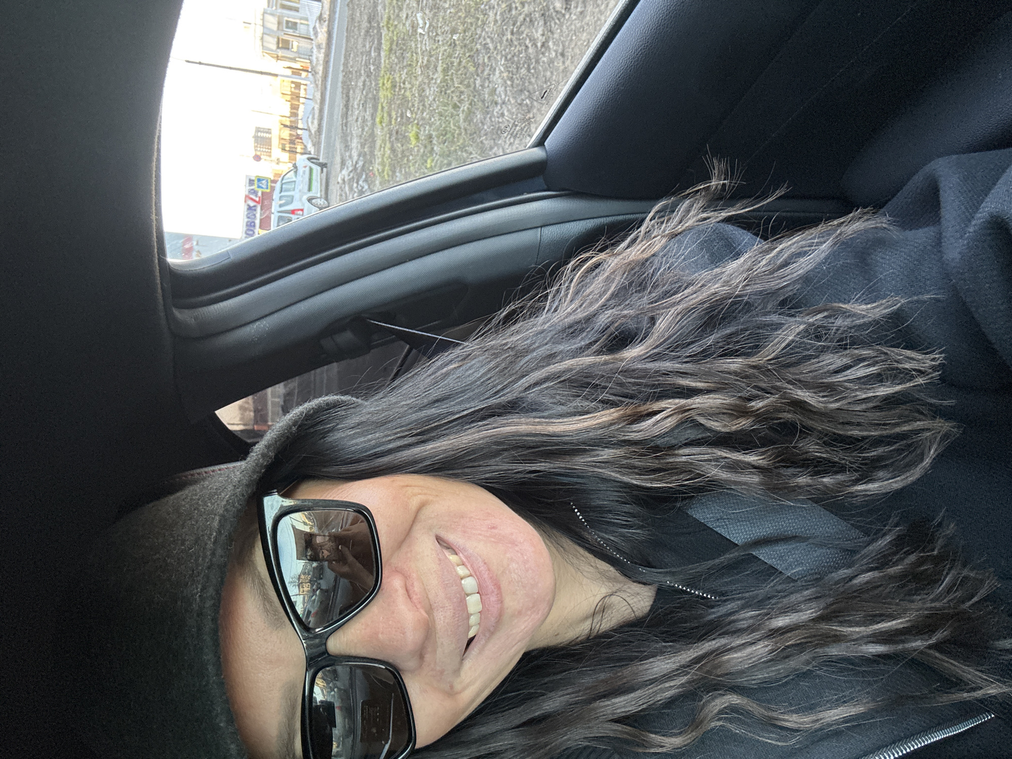 A woman wearing sunglasses and a dark hat is in a car, likely the driver. Her hair is long and wavy with some light highlights. The interior of the car is visible, including the dashboard and door frame. Outside the car window, there is a street scene with buildings and a vehicle marked "Korobka". Based on the Cyrillic lettering, the location is Chaikovsky, Russia. The lighting suggests it is daytime.