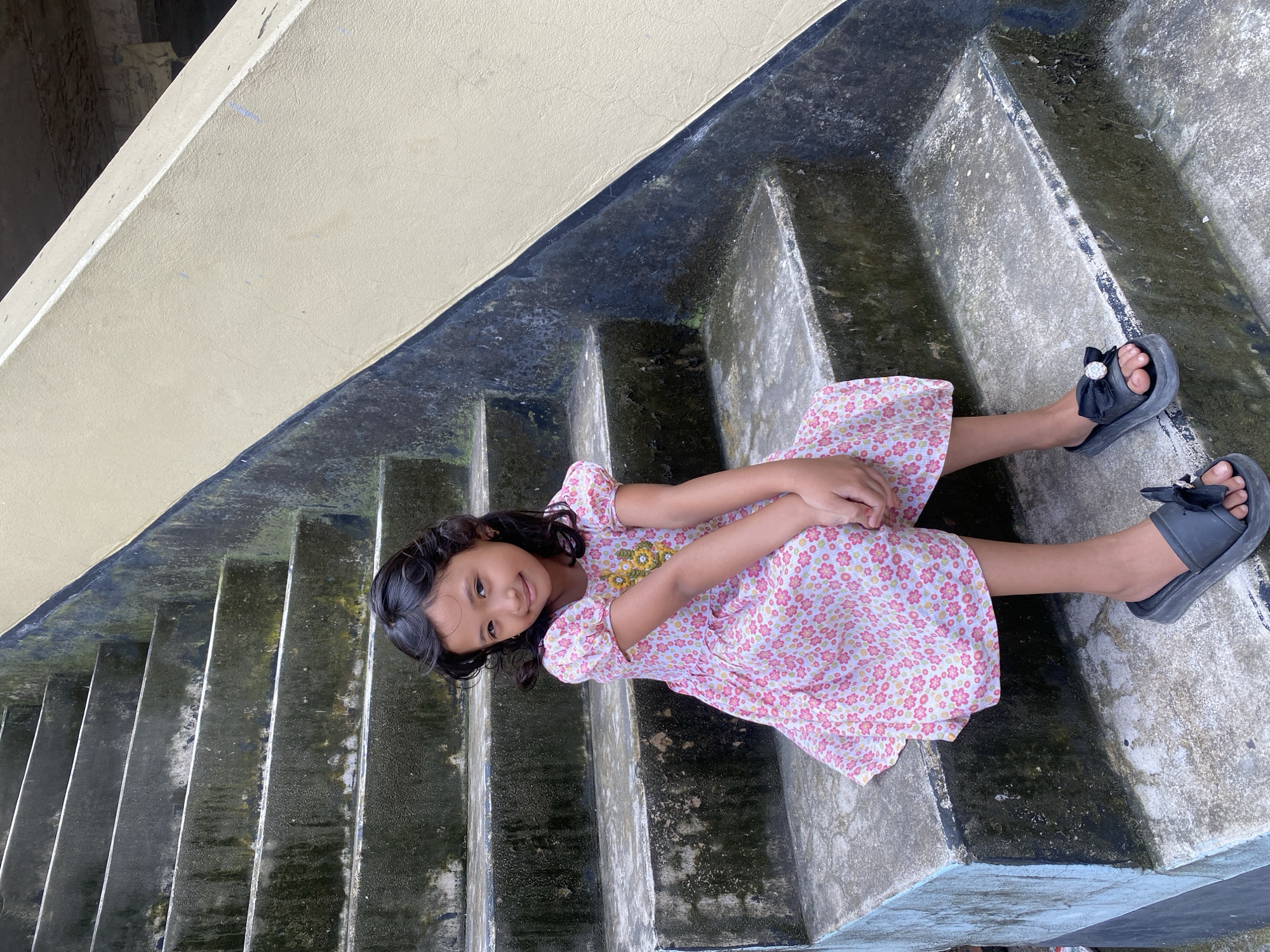 A young girl with dark, curly hair and a pink floral dress sits on concrete steps. She is smiling at the camera, with her hands clasped in her lap. The steps are weathered and have moss growing on them. A yellow wall runs along the side of the stairs. The image appears to be taken outdoors in a residential area, possibly in the city of Dhaka, Bangladesh.