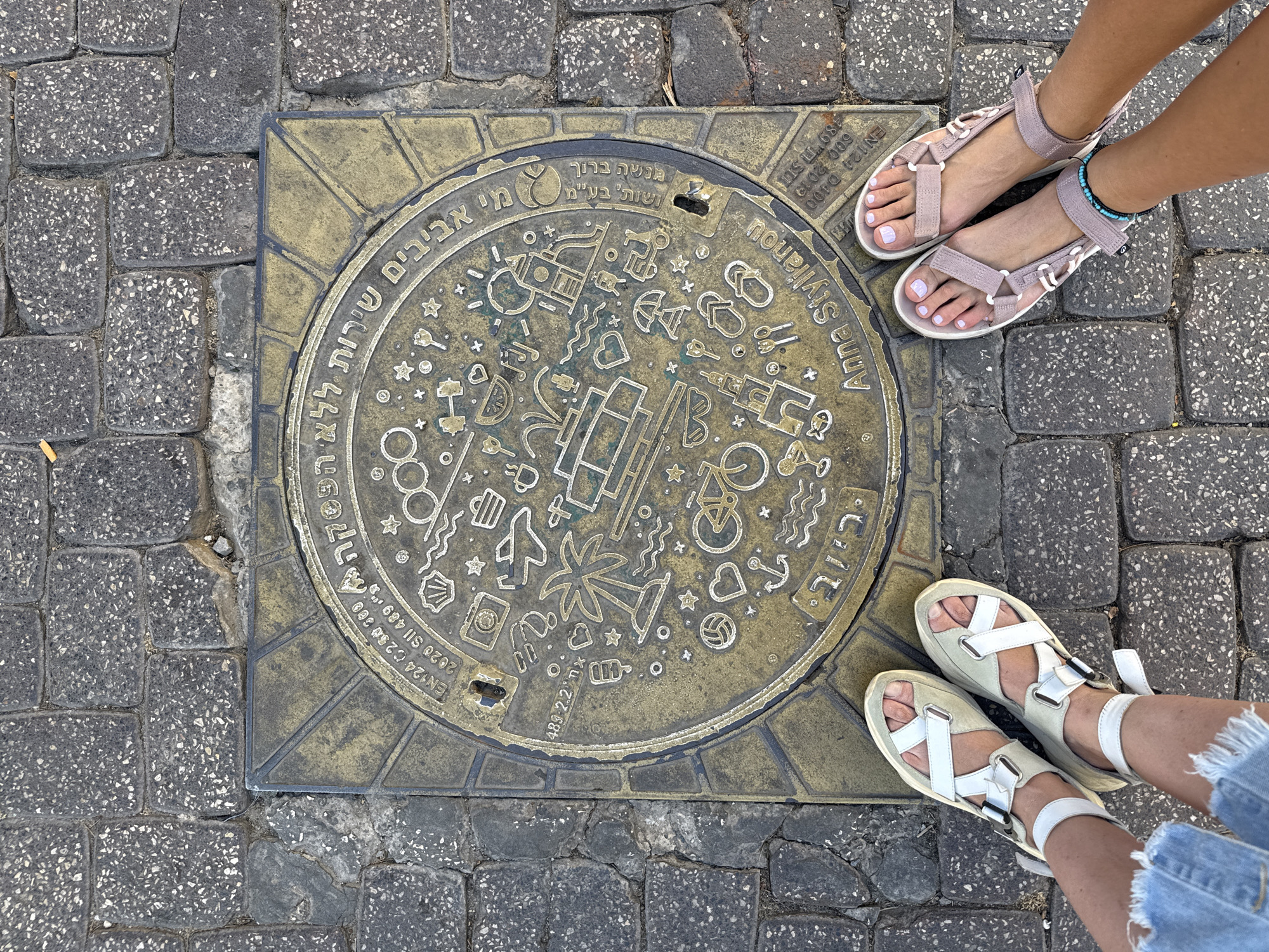 The image shows a close-up, top-down view of two pairs of feet wearing sandals, standing on a cobblestone street next to a decorative manhole cover. The manhole cover is circular and made of metal, with an embossed design featuring various symbols like a palm tree, a bicycle, a heart, an anchor, a volleyball, and other abstract shapes. Hebrew text is inscribed around the rim of the manhole cover, along with English text that includes "Anna Stylianou" and technical specifications like "EN124 C250 300."
One person is wearing light pink, strappy sandals and has a turquoise and black beaded anklet on their left leg. Their toenails are painted a light lilac color. The other person is wearing white and beige chunky sandals with multiple straps. They are also wearing denim shorts with a frayed hem.
The scene appears to be set in a city, possibly Tel Aviv, Israel, as indicated by the Hebrew text and the "Anna Stylianou" inscription which might be a dedication. The lighting suggests it is daytime, and the overall impression is casual and summery, given the footwear and the decorative, beach-themed symbols on the manhole cover. The focus is on the feet and the manhole cover, creating an urban detail shot.