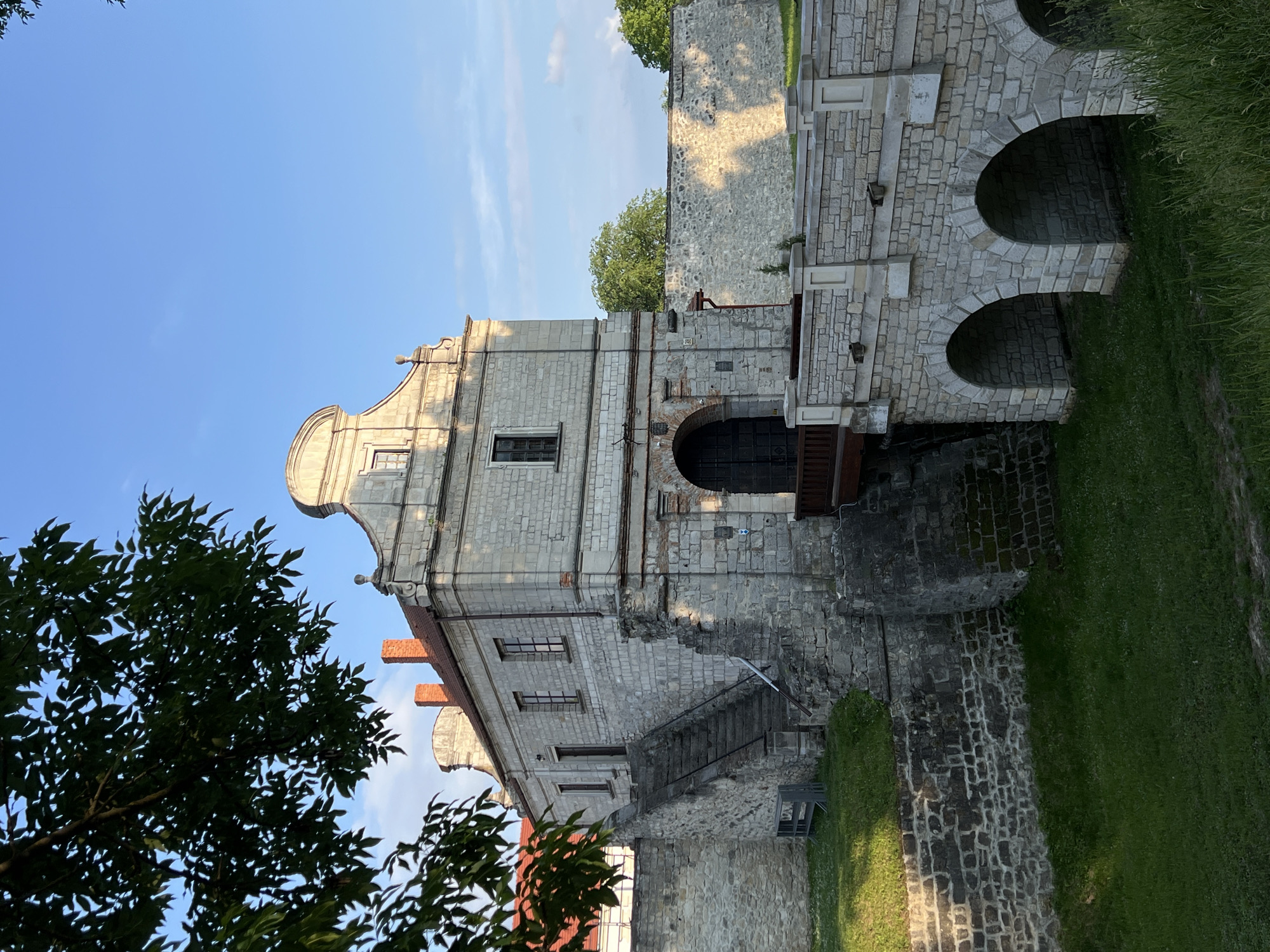 This image depicts a historic stone castle or fortified palace, situated in Zbarazh, Ukraine, under a clear blue sky. The main structure is built from light-colored, roughly cut stone blocks, giving it a robust and aged appearance. A notable architectural detail is the ornate, curved gable atop one section of the building, characteristic of Baroque design. The building features multiple windows with dark frames and a reddish-brown tiled roof with visible chimneys.
To the right of the central building, a stone structure with two prominent arched passages extends, likely serving as a bridge or part of an entrance to the complex. Extensive stone walls, constructed from similar materials, form part of the fortifications or retaining structures around the site.
The ground surrounding the castle is covered in vibrant green grass, suggesting a well-maintained historical site. A large tree with dark green foliage partially obscures the left side of the building and the sky, while other trees are visible in the background. The sky is a bright, clear blue with minimal clouds. The lighting suggests a sunny day, possibly late afternoon or early morning, with strong sunlight highlighting the stone and casting clear shadows. The scene is devoid of people or specific activities, focusing on the architectural grandeur and historical significance of the location. No visible text is present.