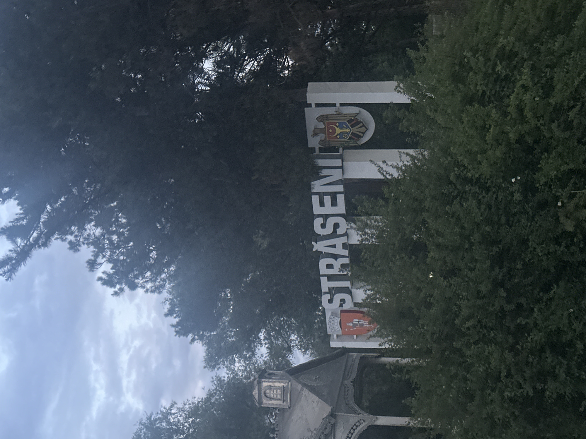 An exterior shot captures a welcome sign for the city of Strășeni, Moldova, under an overcast sky. The sign is large and white with dark lettering spelling out "STRĂȘENI." The city's coat of arms is displayed on the left side of the sign, featuring a red shield with a knight. On the right side of the sign, the national coat of arms of Moldova is visible, a double-headed eagle holding a shield with aurochs head. To the left of the sign is an elaborately decorated, traditional wooden structure with a peaked roof and a small arched window. Dense green foliage, likely bushes and trees, partially obscures the lower portion of the sign and the wooden structure. The overall scene suggests a city entrance or roadside marker.