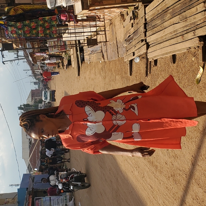 This image captures a wide-angle view of an unpaved, reddish-brown dirt street in Eziobodo, Nigeria. In the immediate foreground, a woman with light brown skin and braided hair styled in a high ponytail walks towards the viewer's left, her head turned to her left, gazing forward. She wears a knee-length, orange button-down shirt-dress featuring a prominent white graphic of Mickey and Minnie Mouse kissing. Her left hand holds a dark-colored mobile phone.
In the mid-ground on the left side of the street, a man on a black motorcycle (okada) rides towards the camera, followed by several pedestrians. The left side of the street is lined with commercial buildings, some with metal roofs; one building features a partial blue facade and a sign reading "MBG." On the right side of the street, a series of informal market stalls and shops with corrugated metal roofs extend into the background. Wooden planks are laid on the ground in front of these stalls, serving as makeshift walkways. Merchandise, including patterned fabrics, hangs from one stall, and a partial sign for "SUNSHINE Resort" is visible. Further in the background, a light-colored vehicle is observed driving away, and additional pedestrians are present. Utility poles with overhead wires are visible along both sides of the street. The sky is overcast.