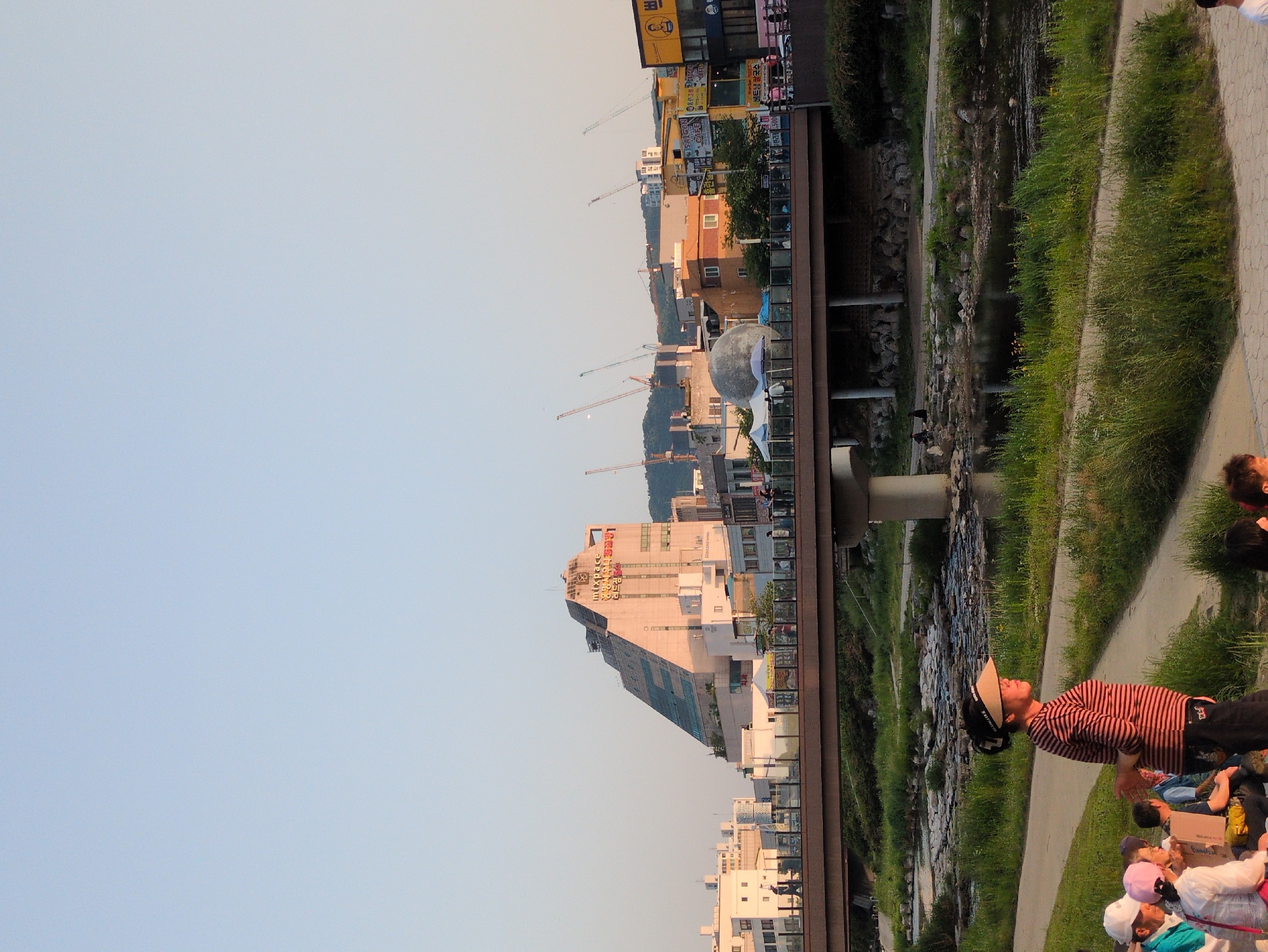 The image captures a scene in Daejeon, South Korea, with a mix of urban and natural elements. In the foreground, people are present, including a person wearing a striped shirt and a hat standing on a paved path. Further along the path, there is a group of people, some of whom are wearing hats. Adjacent to the path is a waterway, bordered by greenery. The background showcases a cityscape with various buildings, including a tall, modern structure with a sloping design and cranes in the distance, suggesting ongoing construction. Other urban elements include a bridge structure spanning the waterway and commercial buildings. The sky above is clear and light blue.
