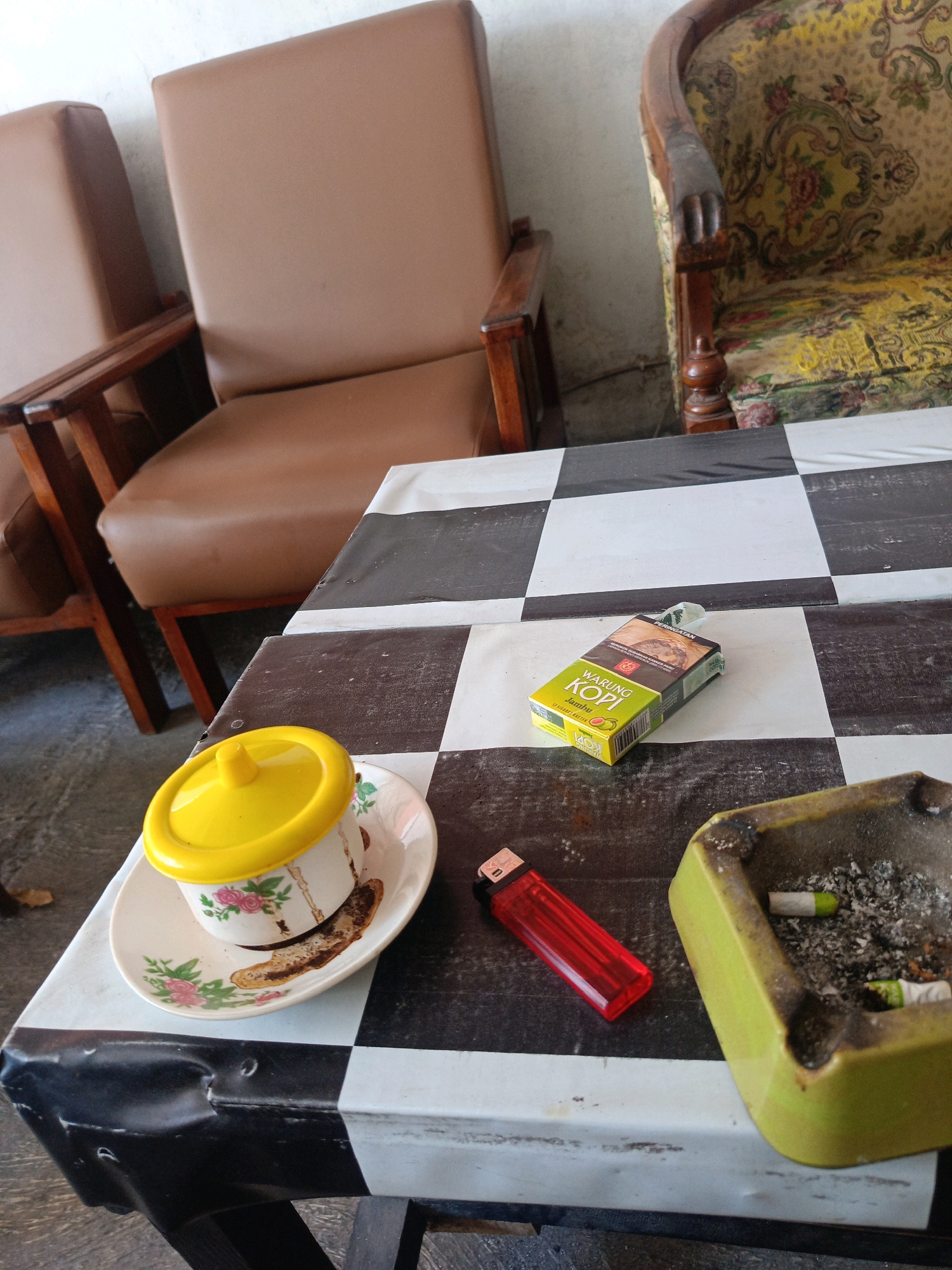 The image depicts a scene on a table in what appears to be an outdoor or semi-outdoor seating area in Candirejo, Indonesia. The table is covered with a black and white checkered cloth. On the table are several objects: a pack of "WARUNG KOPI JAMBU" cigarettes, a red plastic lighter, and a light green ashtray containing cigarette butts and ash. To the left of these items is a white cup and saucer with floral decorations, topped with a bright yellow lid. In the background, two chairs are visible: one is a brown leather armchair, and the other is an older armchair upholstered with a yellow floral fabric. The overall impression is of a resting spot, possibly a cafe or a home patio, where someone has been smoking and drinking.