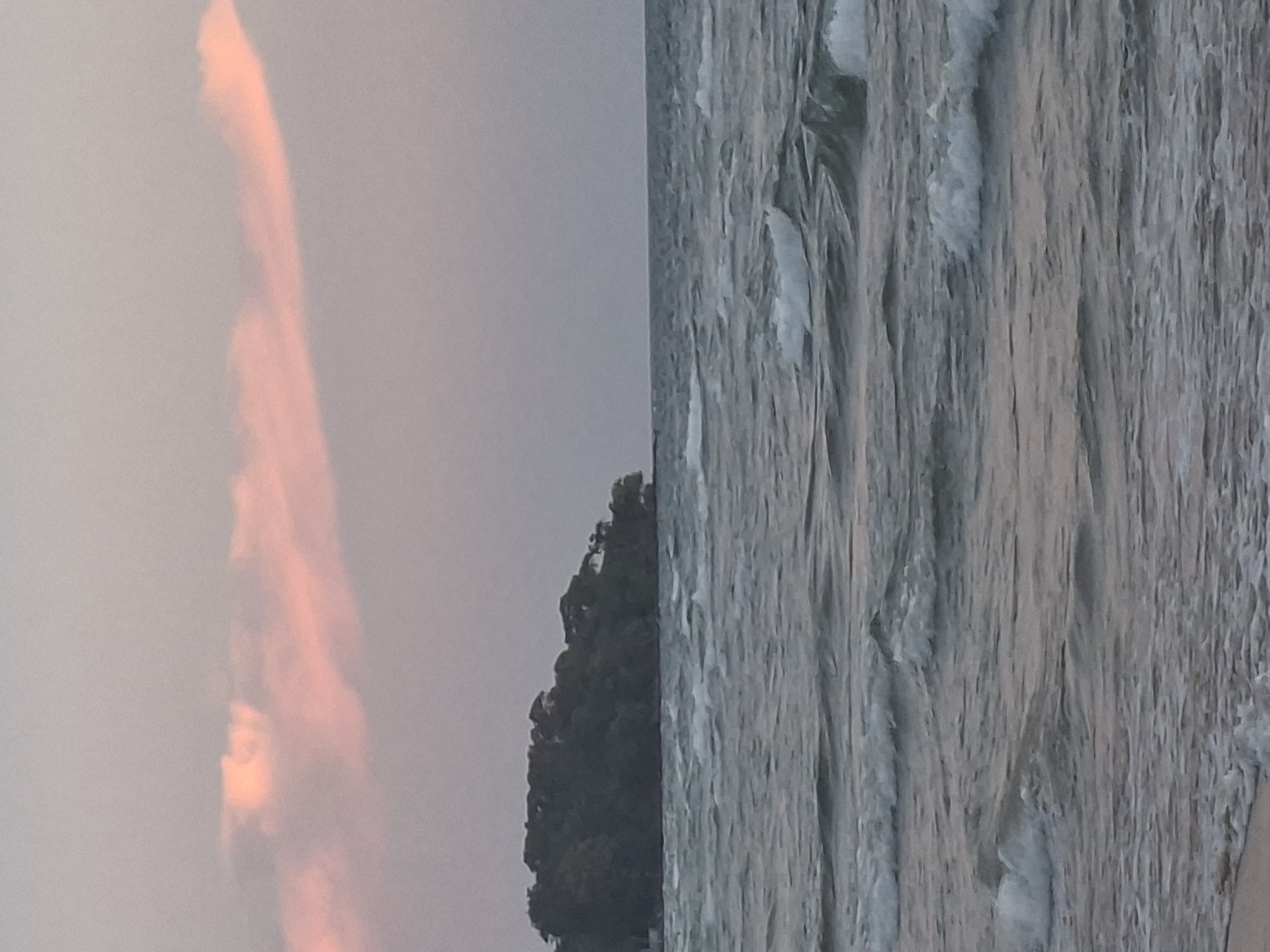 An island covered in dense green foliage juts out from the ocean, with a sliver of sand visible in the foreground. The sea is choppy, with waves rolling in, and a soft pink cloud drifts across the pale, overcast sky. The scene is likely from the southern coast of Phú Quốc, Vietnam, possibly near An Thoi archipelago, as islands are a common feature of this region.