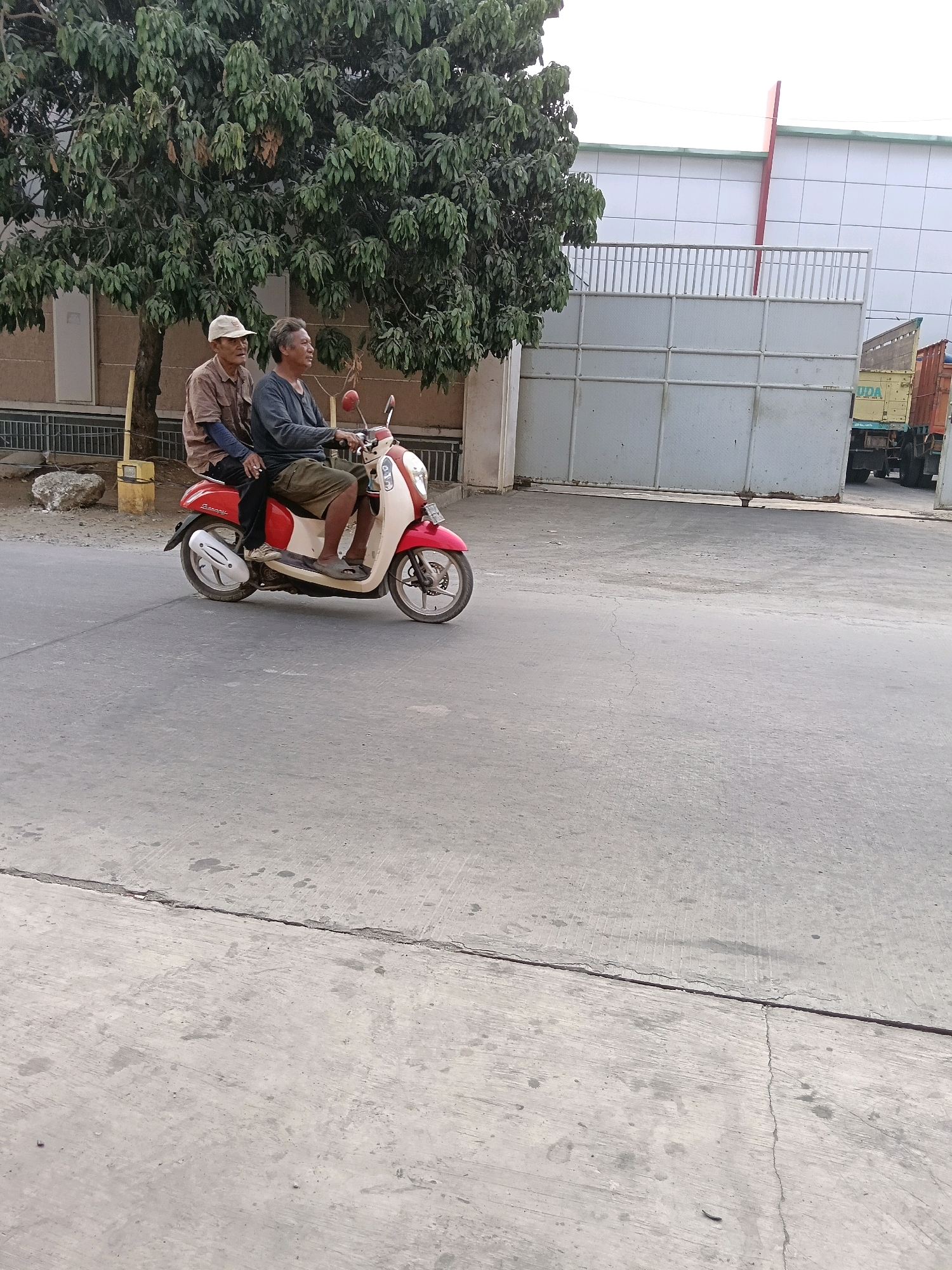 Two men are riding on a red and white scooter on a concrete road. The man in front is wearing a dark long-sleeved shirt and olive green shorts, while the man behind him is wearing a light brown short-sleeved shirt and a tan baseball cap. They are positioned in the middle of the road, which is cracked and appears worn. In the background, there is a large tree with dense green foliage on the left, and a building with a white tiled facade and a large grey gate on the right. A truck is partially visible behind the gate. The overall scene suggests an outdoor, possibly industrial or commercial area in Belimbing, Indonesia, during daylight hours.