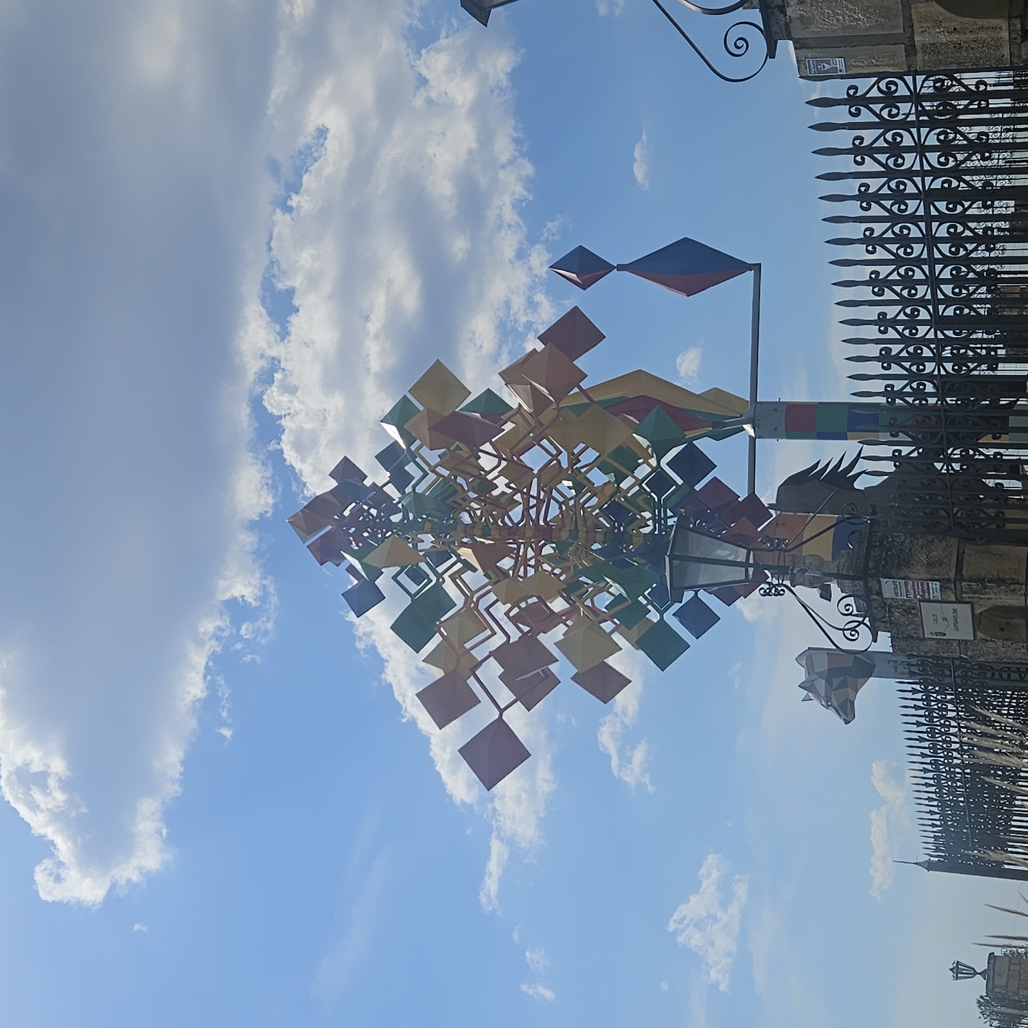 The image shows a low-angle view of a public art installation in Algora, Spain, set against a blue sky with scattered clouds. The installation features numerous colorful diamond-shaped metal plates attached to a complex framework. It is located near a wrought iron fence and gate, which are partially visible on the right side of the frame. A stylized rooster sculpture is also present. A street sign indicating "Calle de Cataviento" can be seen attached to a stone structure near the gate.