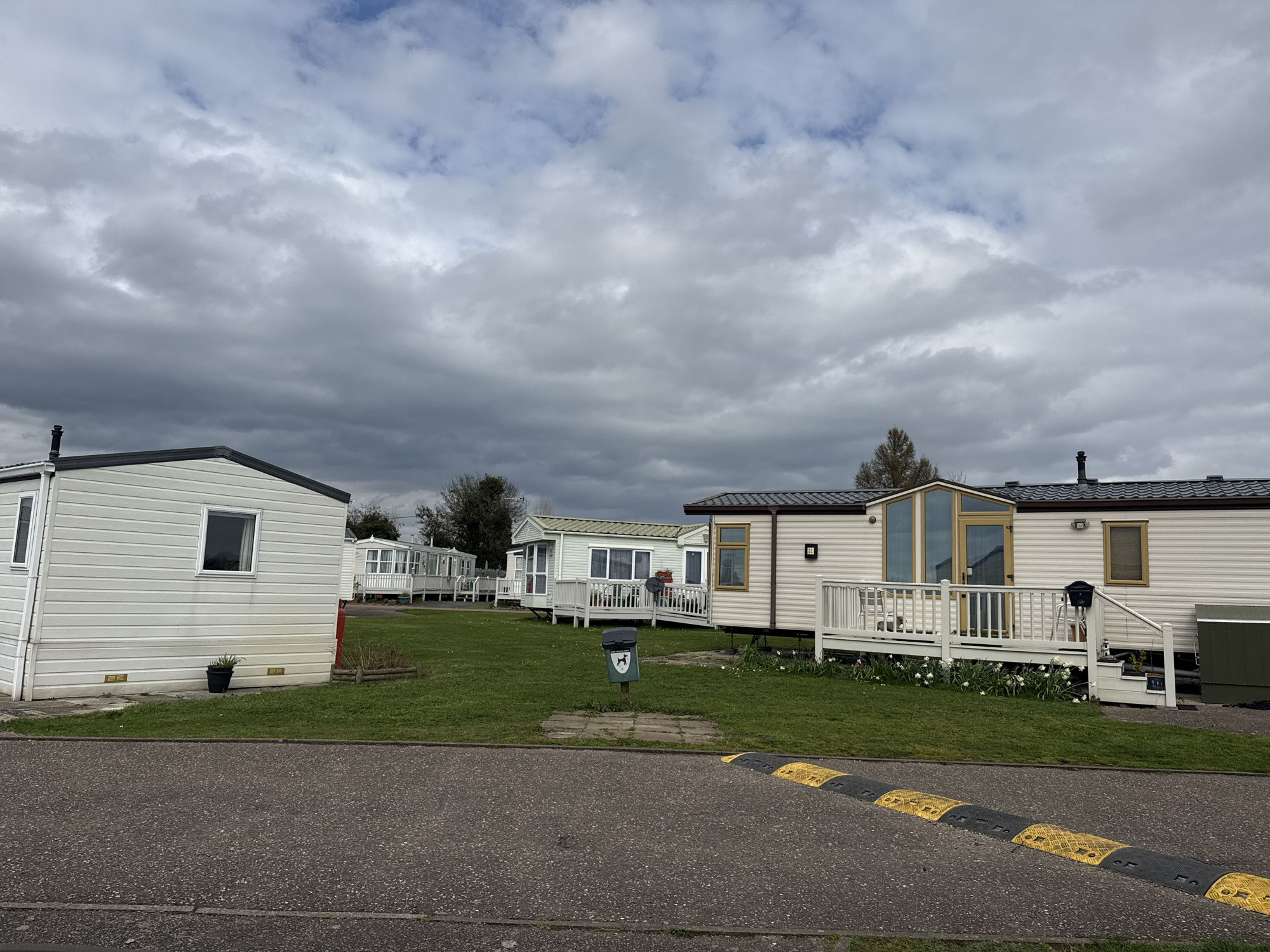 The image depicts a caravan park in Great Yarmouth, United Kingdom on a cloudy day. Multiple white static caravans with dark trim are visible, arranged in rows on green lawns. Some have small decks with white railings. A gray asphalt road with a black and yellow speed bump runs in the foreground. There is a green dog waste bin near the road. The sky is overcast with heavy gray clouds.