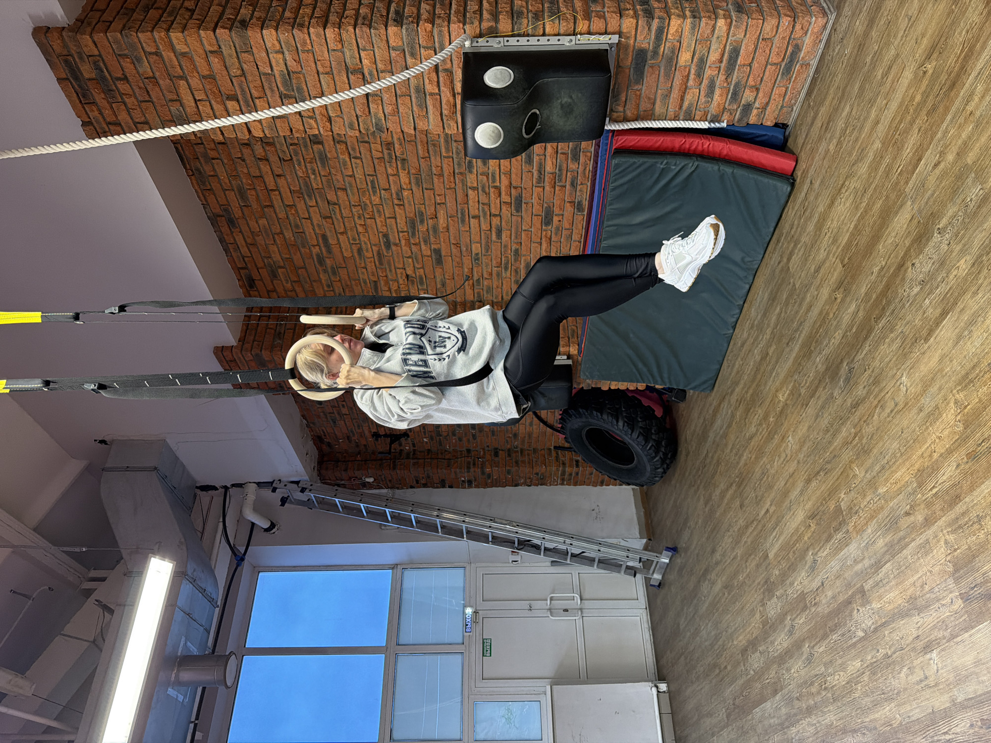 The image shows a woman exercising in a gym. She is suspended from gymnastic rings. She is wearing black leggings, a grey sweatshirt, and white sneakers. Behind her are brick walls, a tire, and mats. A white rope runs along the ceiling. In the background, there is a window, a door, and a ladder. Based on available data, the location is the city of Chaikovsky, Russia.