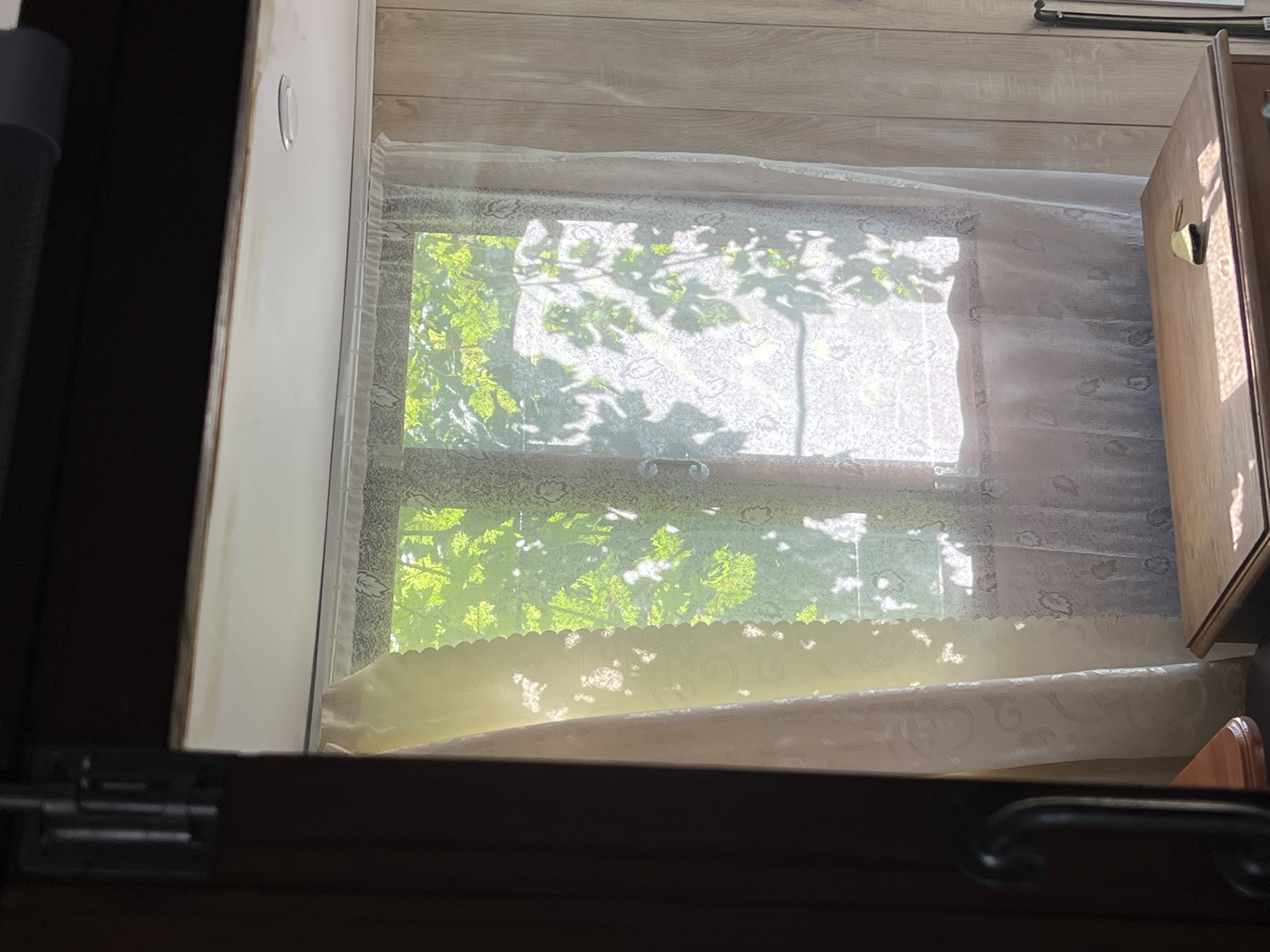 The image depicts an interior scene looking out a window. The window is covered by sheer lace curtains with a floral pattern, and a lighter, yellow-tinted sheer curtain is partially drawn to the left. Sunlight streams through the window, casting dappled shadows of green foliage on the curtains. Outside the window, vibrant green leaves are visible, suggesting the presence of trees or other plants.
In the foreground, a wooden table or dresser is visible, with a small, yellow object, possibly a key or a tag, resting on its surface. The table exhibits visible wood grain and is positioned in front of the window. To the left, a dark wooden door frame or furniture piece is partially visible, with a dark, curved handle. The wall to the right of the window is paneled with light-colored wood.
There are no people visible in the image. The overall atmosphere is peaceful and bathed in natural light. The scene appears to be a domestic interior, possibly a room in a house or cabin. The mention of Khryashchevka, Russia, places this scene geographically.