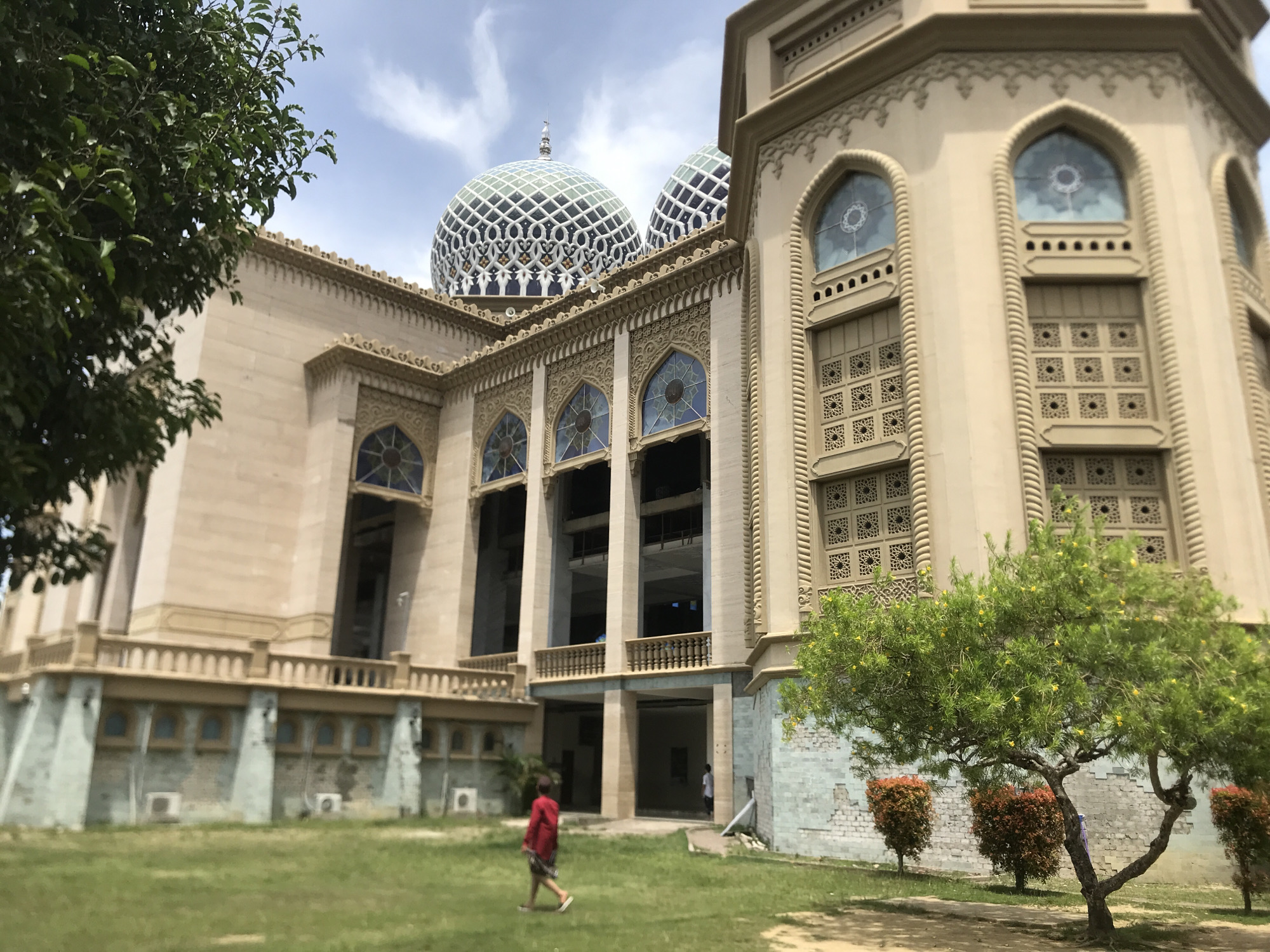 The image depicts a large, ornate mosque in Lhokseumawe, Indonesia. The building is constructed from light-colored stone and features multiple domes with blue and white patterned exteriors. The architecture includes tall, arched windows, decorative moldings, and intricately patterned screens. A person wearing a red shirt is walking across a grassy lawn in front of the mosque. A small tree with yellow flowers stands to the right of the building. The sky is partly cloudy.