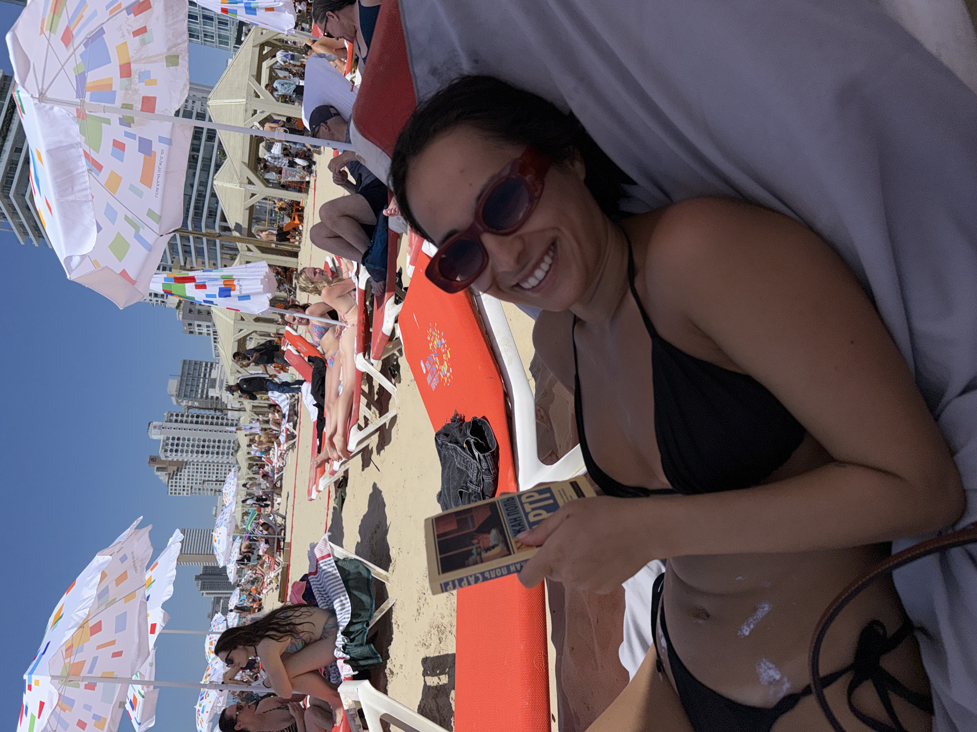 On a sunny day at a Tel-Aviv beach, a woman lies on a lounge chair, wearing sunglasses and a black bikini. She holds a book open in her hands, looking at the camera and smiling. Sunscreen is visible on her stomach. In the background, other beachgoers recline on chairs under colorful umbrellas, with tall buildings lining the horizon. The sand is light brown, and the sky is clear blue.