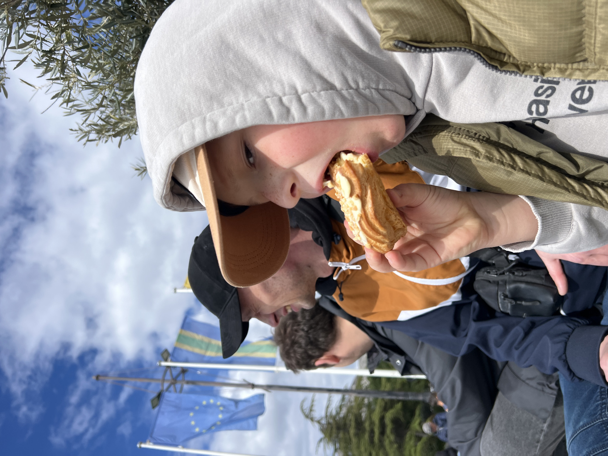 A low-angle, medium shot depicts a child in a grey hooded sweatshirt, with a brown baseball cap underneath, eating a pastry. The child is also wearing a green vest. Behind the child is a man wearing a black baseball cap and a yellow and navy jacket. Further in the background are flags, including a European Union flag, and trees. The scene is outdoors on a sunny day with a blue sky and white clouds. Given the location and context, this is likely taken in Bar, Montenegro.