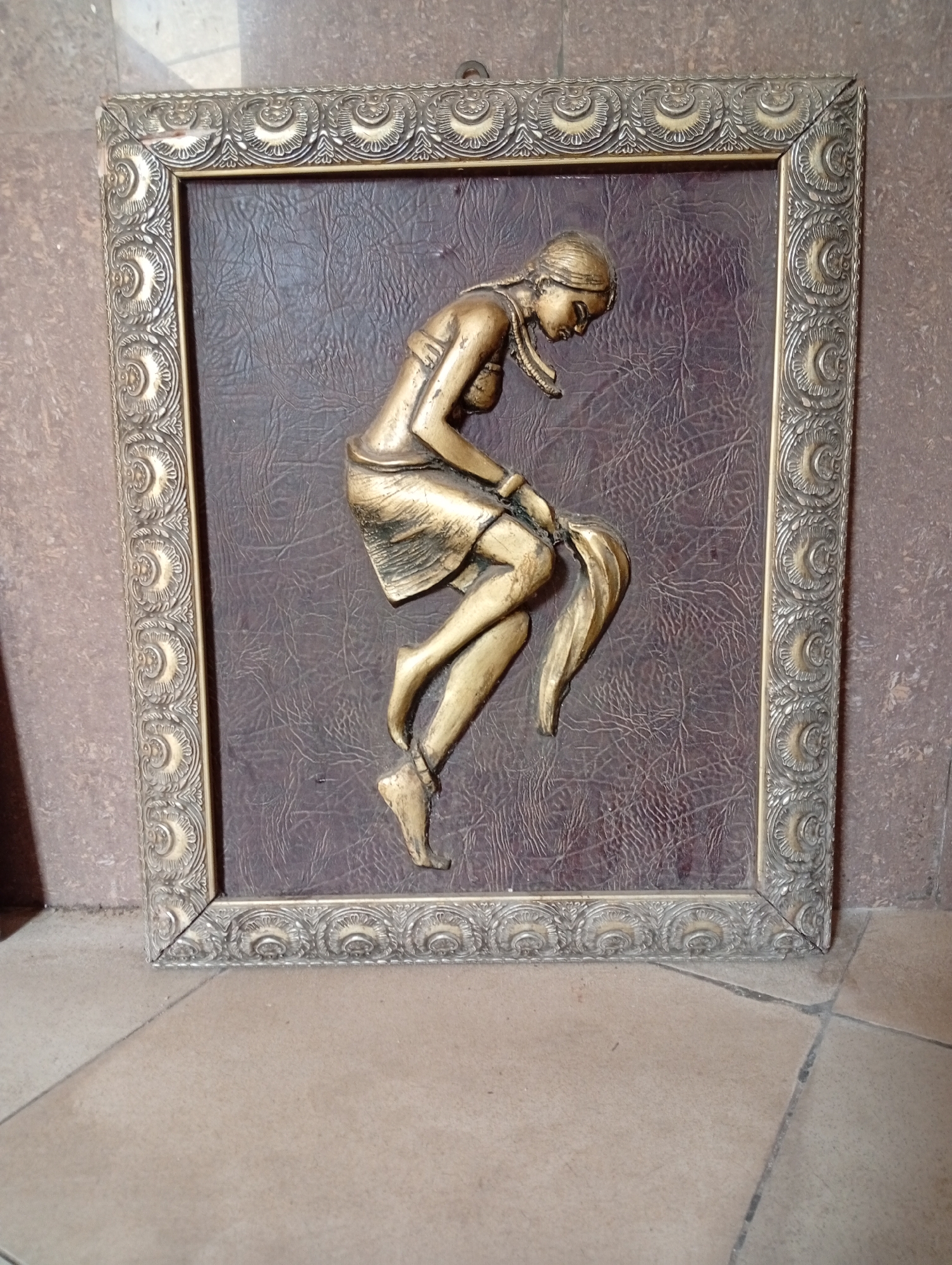 The image depicts a framed three-dimensional artwork. The frame is ornate, with repeating decorative elements in a metallic gold or bronze color. The artwork within the frame features a raised figure of a woman, possibly made of metal or a similar material, also finished in a metallic gold or bronze tone. The woman is depicted in a seated or squatting position, possibly with fabric or a garment hanging from her hand or waist. The background of the artwork is a textured, possibly leather-like material, in a dark brownish-purple color. The artwork is leaning against a wall in a location with tiled flooring. The location is unknown. No person in the city of Adankolo, Nigeria, is visible.