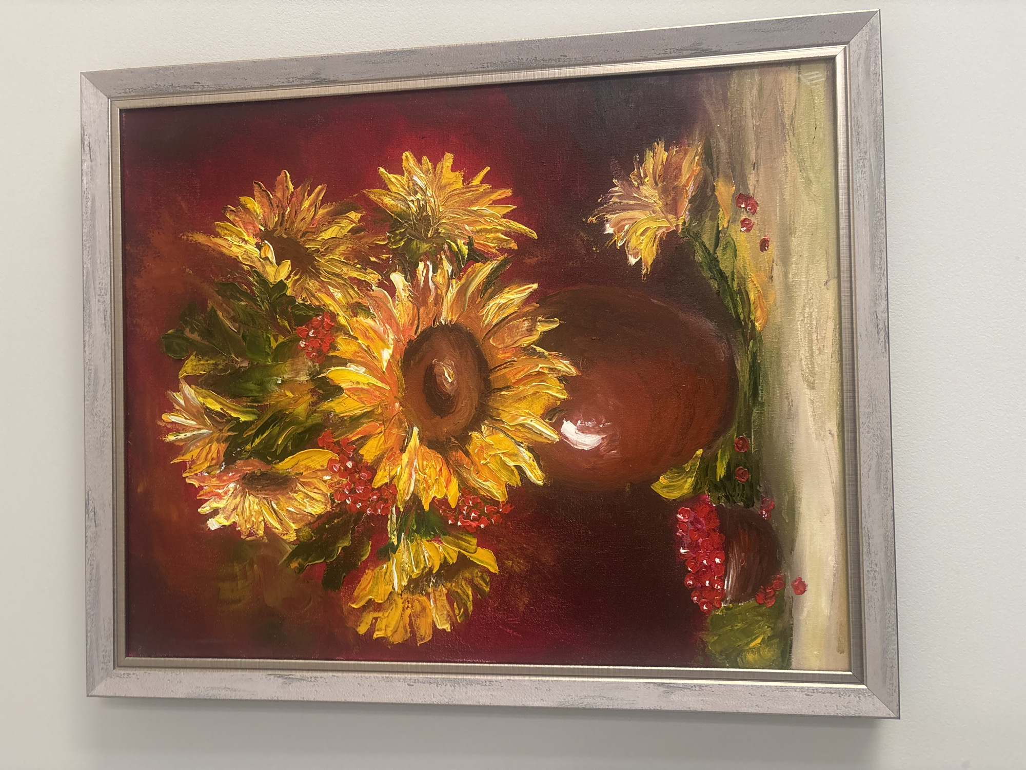 A framed oil painting depicts a still life of sunflowers in a vase, accompanied by berries. The sunflowers are predominantly yellow and orange, with thick, textured brushstrokes that emphasize their petals. Red berries are scattered throughout the arrangement, some in a small, dark bowl to the left of the vase, and others loose on the surface below. The vase is a deep brown, with a glossy finish. The background is a rich, dark red, transitioning to lighter tones in some areas, creating depth. A single fallen sunflower head with yellow petals rests on the surface to the right of the vase. The painting is framed in a light gray, textured wooden frame. The artwork is hung on a white wall. The provided context mentions the city of Pereiaslavske, Ukraine, but the image itself does not contain any visual cues to confirm this specific location.