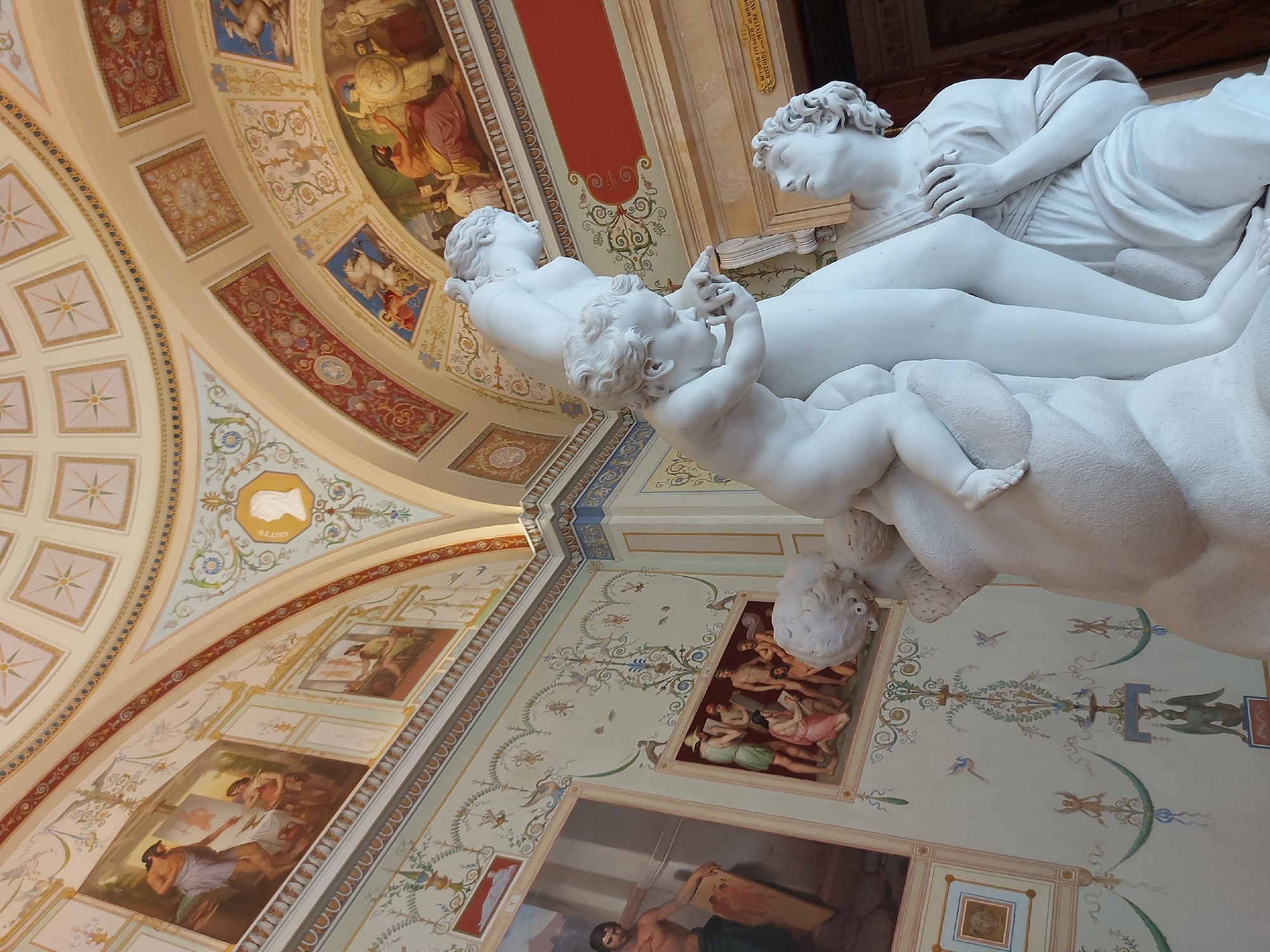 The image shows a marble sculpture of a woman, a baby, and a man, viewed from below, with decorative painted ceilings and walls as the backdrop. The location is the Hermitage Museum in Saint Petersburg, Russia. The sculpture depicts a reclining woman holding an infant, with a man standing and leaning over them. The ceilings are elaborately decorated with geometric patterns, floral motifs, and painted scenes in rectangular frames. The walls are also adorned with paintings in ornate frames.