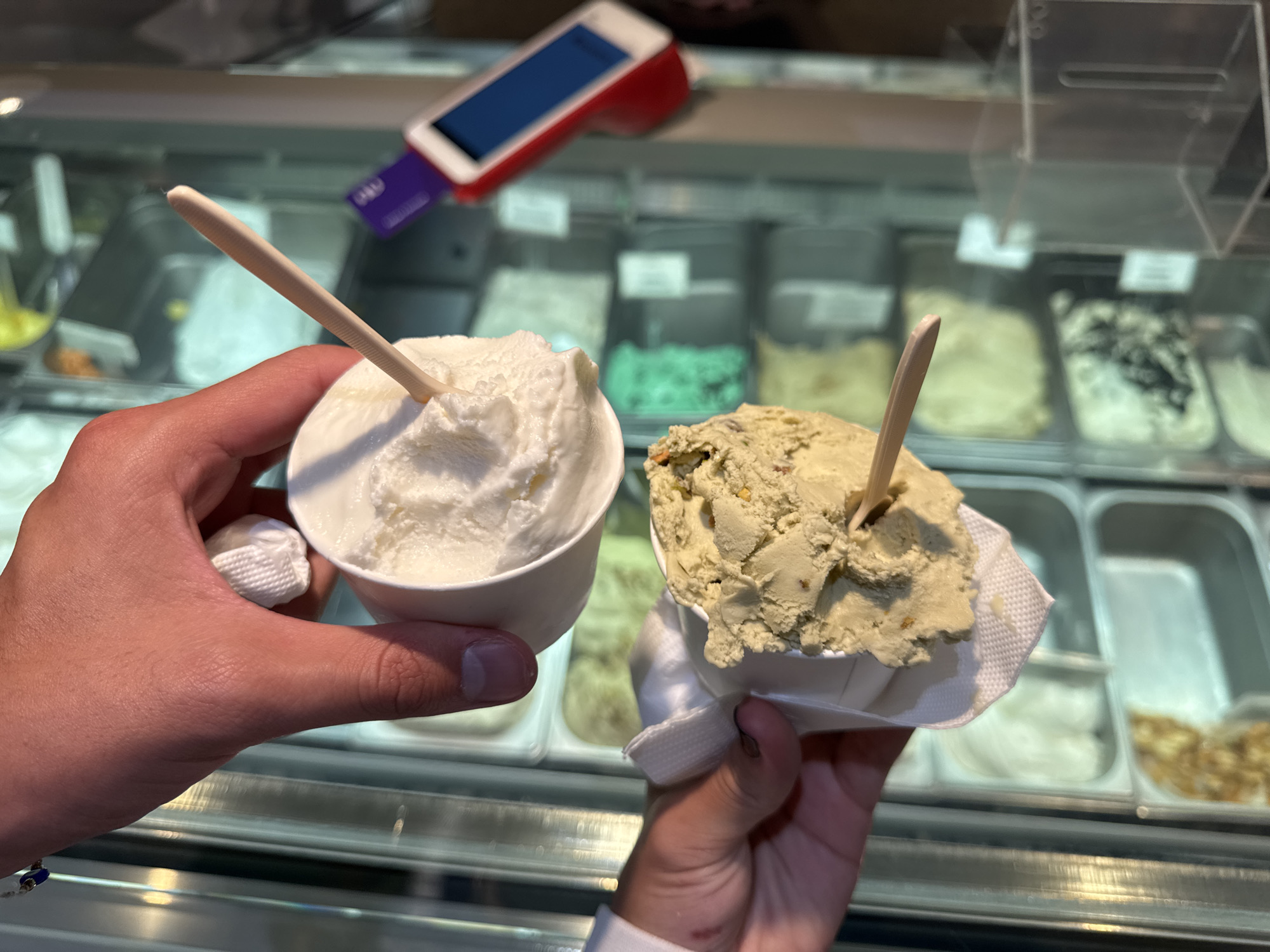 The image shows two hands holding cups of gelato with wooden sticks. The cup in the left hand contains white gelato. The cup in the right hand contains pistachio gelato. Behind the hands and gelato is a glass display case containing various flavors of gelato. A red and white card reader sits on top of the display case. The location is likely a gelato shop in Mexico City, Mexico.