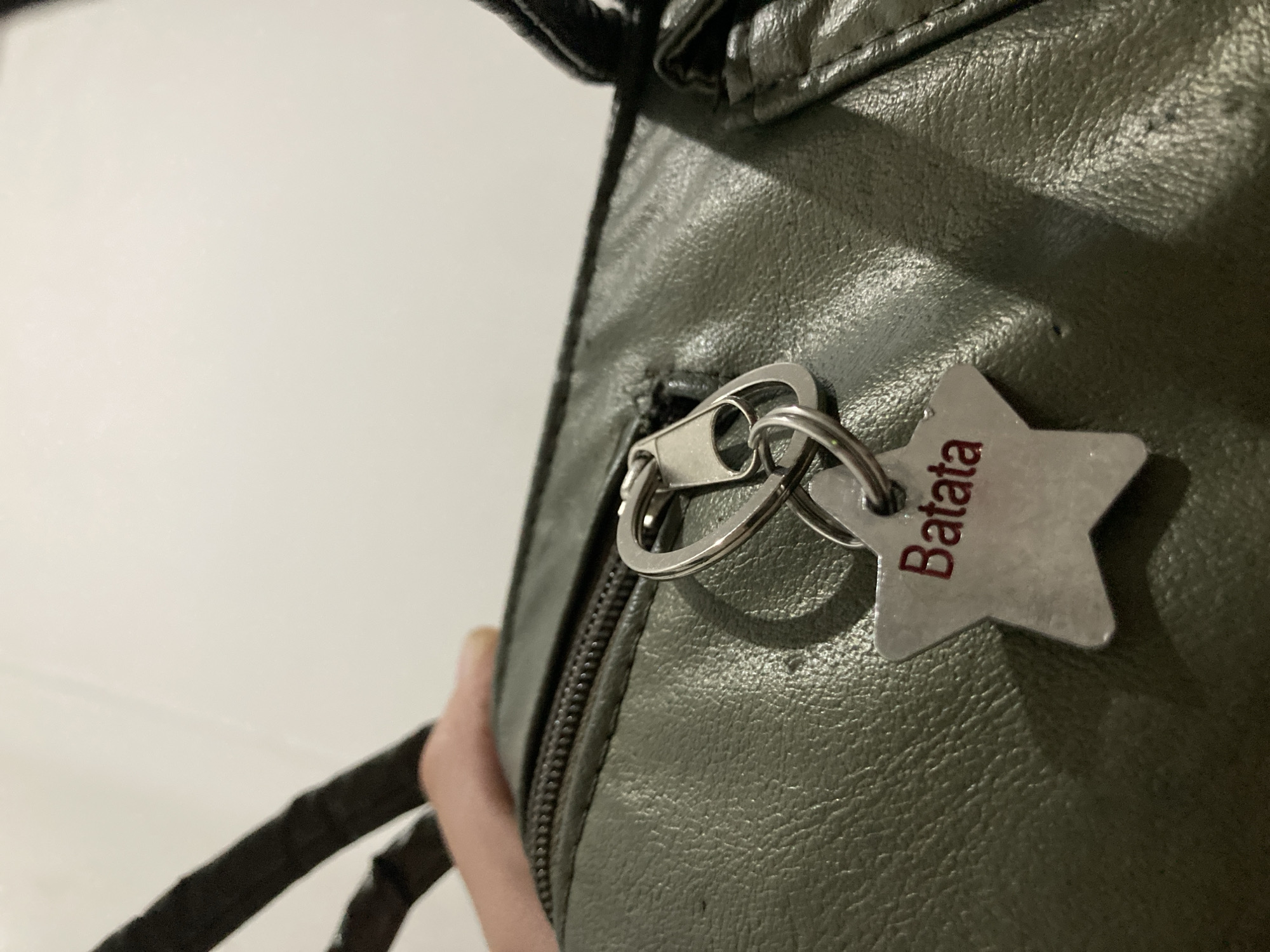 A close-up shot shows a portion of a green leather bag. A zipper with a silver pull is visible on the bag. Attached to the zipper pull is a silver star-shaped keychain with the word "Batata" inscribed in red letters. The background is a plain, off-white color. A portion of the bag's strap is visible on the left. The location is Unknown, Unknown.