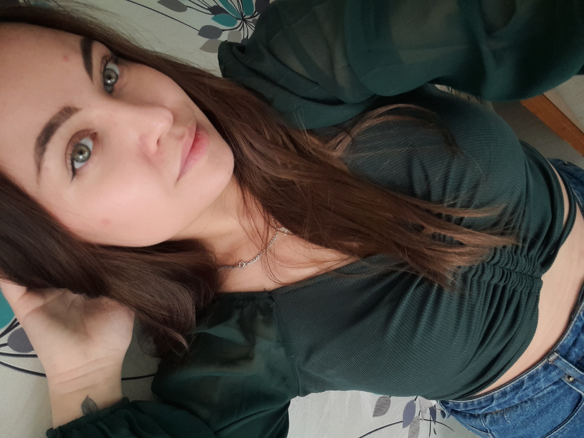 A woman with fair skin, brown hair, and green eyes is seen in a close-up shot. She is wearing a dark green top and blue jeans. A silver necklace is visible around her neck. The background shows a surface with a floral pattern. A small leaf tattoo is visible on her wrist. The location is Unknown, Unknown.