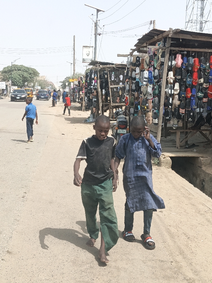The image captures an active street scene in Hotoro South, Nigeria, under clear daylight conditions. In the foreground, two young males walk leftward on a dirt shoulder adjacent to an asphalt road. The boy on the left wears a black t-shirt with white trim and green pants, appearing barefoot. The boy on the right wears a blue plaid tunic over dark pants and black Adidas slide sandals; he holds a dark object to his right ear, consistent with a mobile phone interaction.
To the immediate right, a series of makeshift market stalls line the dirt shoulder. These structures are built with wooden frames and corrugated metal roofs and are densely packed with footwear, predominantly colorful sandals and slides, displayed by hanging. A motorcycle, bearing Nigerian license plate "JGE 671VF," is partially visible parked near these stalls. A concrete drainage ditch runs along the right side of the road.
On the asphalt road, a young male in a blue shirt and dark pants walks away from the camera. Further down the street, another individual in a red shirt walks towards the right. Several vehicles, including a yellow taxi and a black sedan, are present on the road. Multiple utility poles, some with overhead wires, extend into the background; one features a solar panel at its apex. A tree with green foliage is visible on the left side of the road in the mid-distance. Shadows are cast to the left, indicating a sunlit environment.