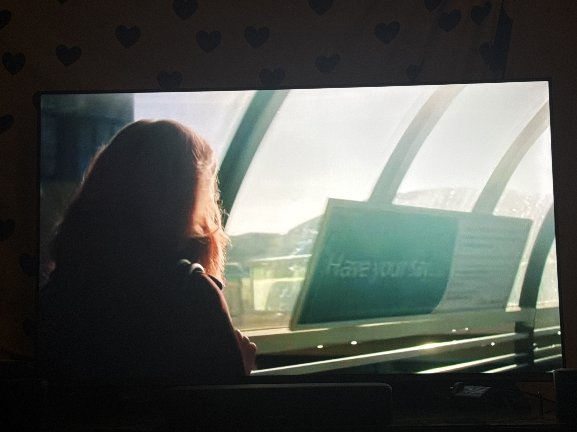 The image shows a television screen displaying a scene from what appears to be a movie or show. In the foreground, a person with reddish-brown hair is seen from the back, with their silhouette largely obscured by shadow. They are looking towards a sign. The sign is a bright teal color with white text that reads "Have your say". The background is blurred and shows a bright, possibly outdoor, environment with what looks like an arched structure. The television is placed in a room, and the wall behind it is decorated with a pattern of black hearts. The bottom of the image shows the top of a dark soundbar or speaker. There is no information in the image that indicates a specific location within the city of Cheltenham, United Kingdom.