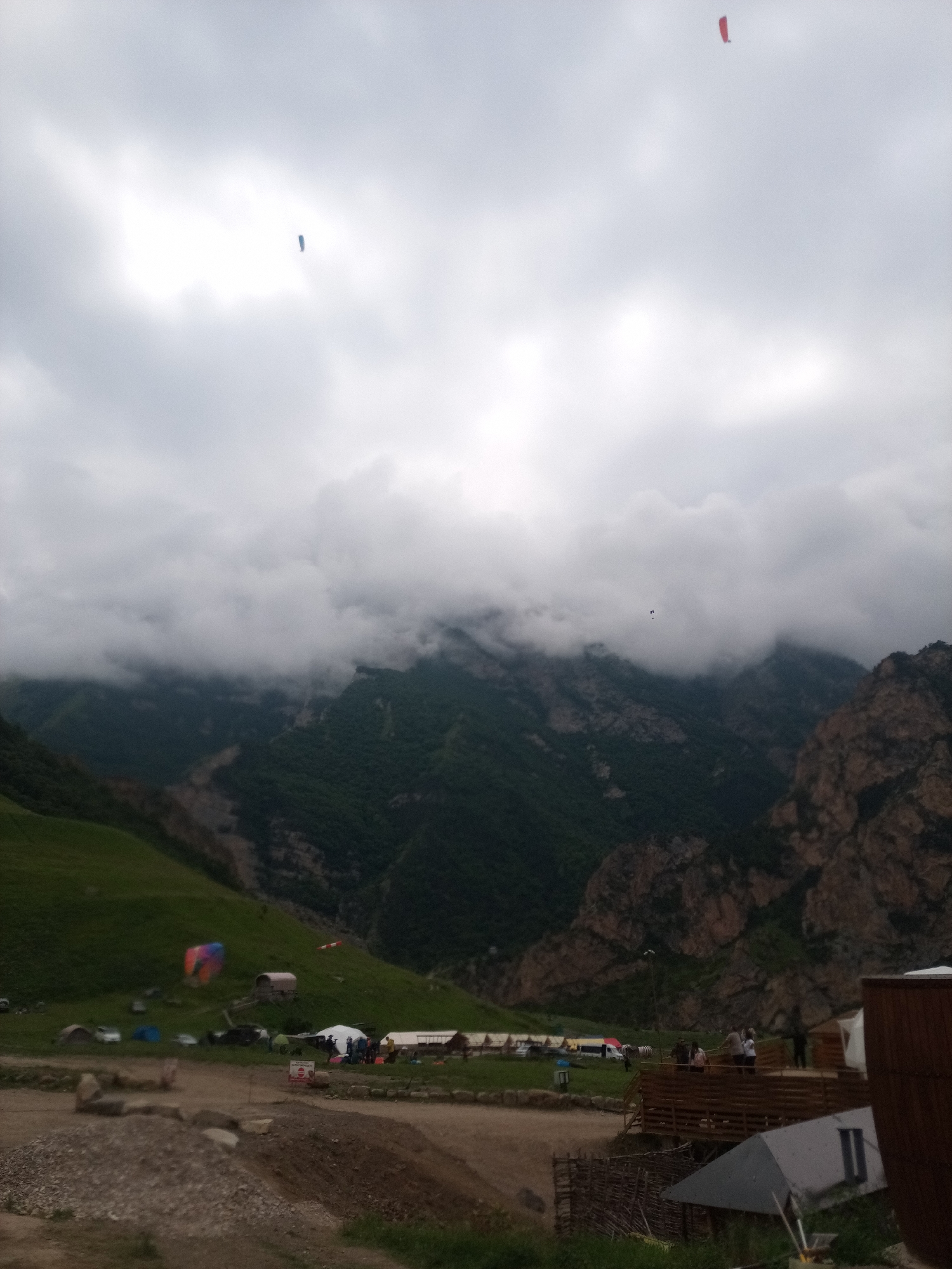 In El'tiubiu, Russia, a wide shot captures a cloudy, overcast sky above a mountainous, green landscape. Two paragliders, one red and one blue, are visible in the sky. Below, a small encampment is set up on a grassy hill, with several tents and a few vehicles. A colorful paraglider is also visible on the ground. Rocky outcrops and forested mountains form the backdrop. The foreground shows a dirt path with some rocks and fencing, suggesting a recreational or adventure tourism area.
