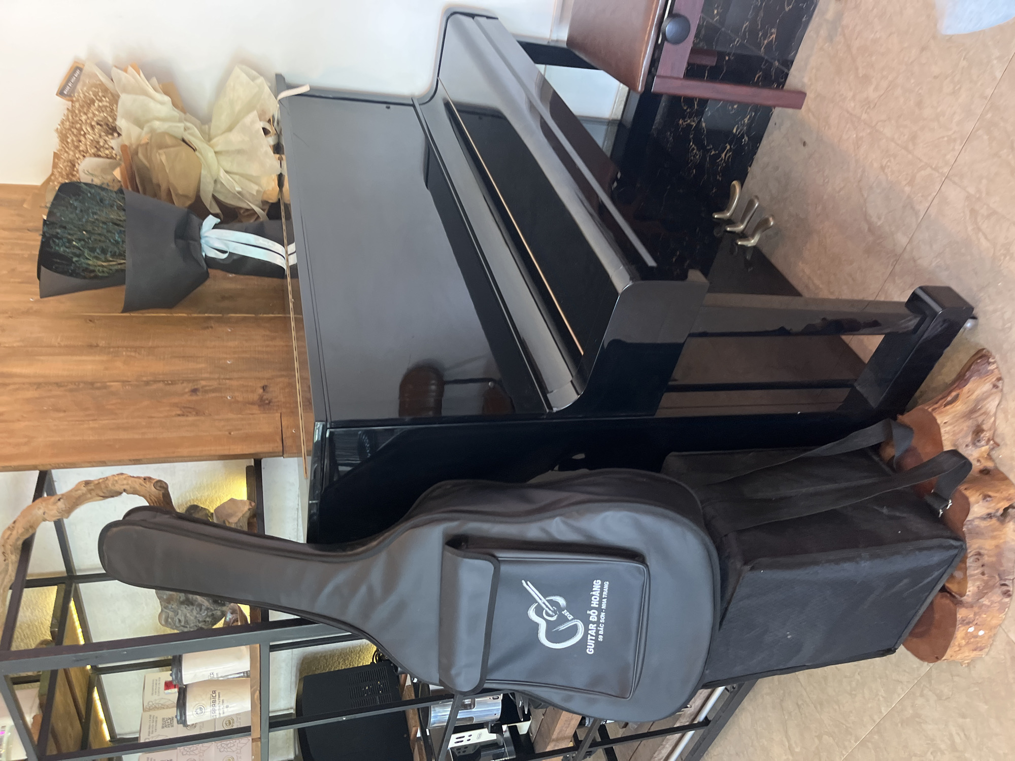 The image depicts a grand piano indoors, with a guitar case and a black bag placed near its base. The location is Bac Nha Trang Ward, Vietnam, as indicated on the guitar case which reads "Guitar Đo Hoang, 59 Bac Son - Nha Trang". The piano is black and reflects ambient light, suggesting a polished surface. The guitar case is gray, with a visible logo and text. The black bag is nondescript and appears to be resting against a decorative wooden piece. The piano is situated near a shelf unit containing various items, including what appears to be coffee products. A small table or stand with a dark marble top is positioned behind the piano.