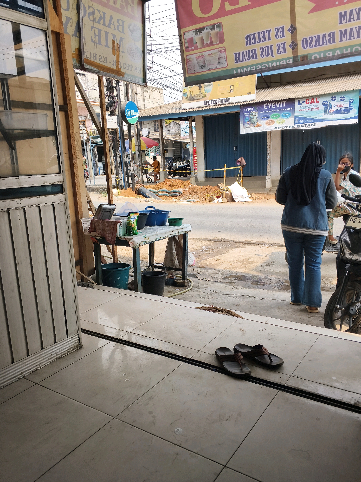 The image depicts a street scene in Sukoharjo III, Indonesia. In the foreground, a tiled floor leads to the entrance of a building, with a pair of brown flip-flops resting on the tiles. To the left, the exterior of a building with vertical siding is visible.
Further into the scene, a table is set up outdoors, covered with a patterned cloth. On the table are various items, including a white plastic basket, several buckets (one green, two blue, and one black), a metal container, and a bottle of liquid soap. A checkered cloth is draped over the basket.
Beyond the table, a street runs perpendicular to the viewer's perspective. On the street, two women are present. One woman, wearing a black hijab and a blue denim jacket with blue jeans, stands facing away from the camera. She is partially obscured by a motorcycle on the right side of the image. Another woman, wearing a patterned top and a black face mask, is also visible, facing the woman in the hijab.
In the background, a street is lined with buildings, some with blue roller shutters. Banners are hung overhead, one of which displays text and images of drinks. Another banner, further down the street, advertises "APOTEK BATAM" and "EYEVIT." Construction or road work is evident in the middle ground, with piles of sand and a large pipe. A man in an orange shirt is operating a wheelbarrow in this area. The overall environment suggests a commercial area with ongoing activity.