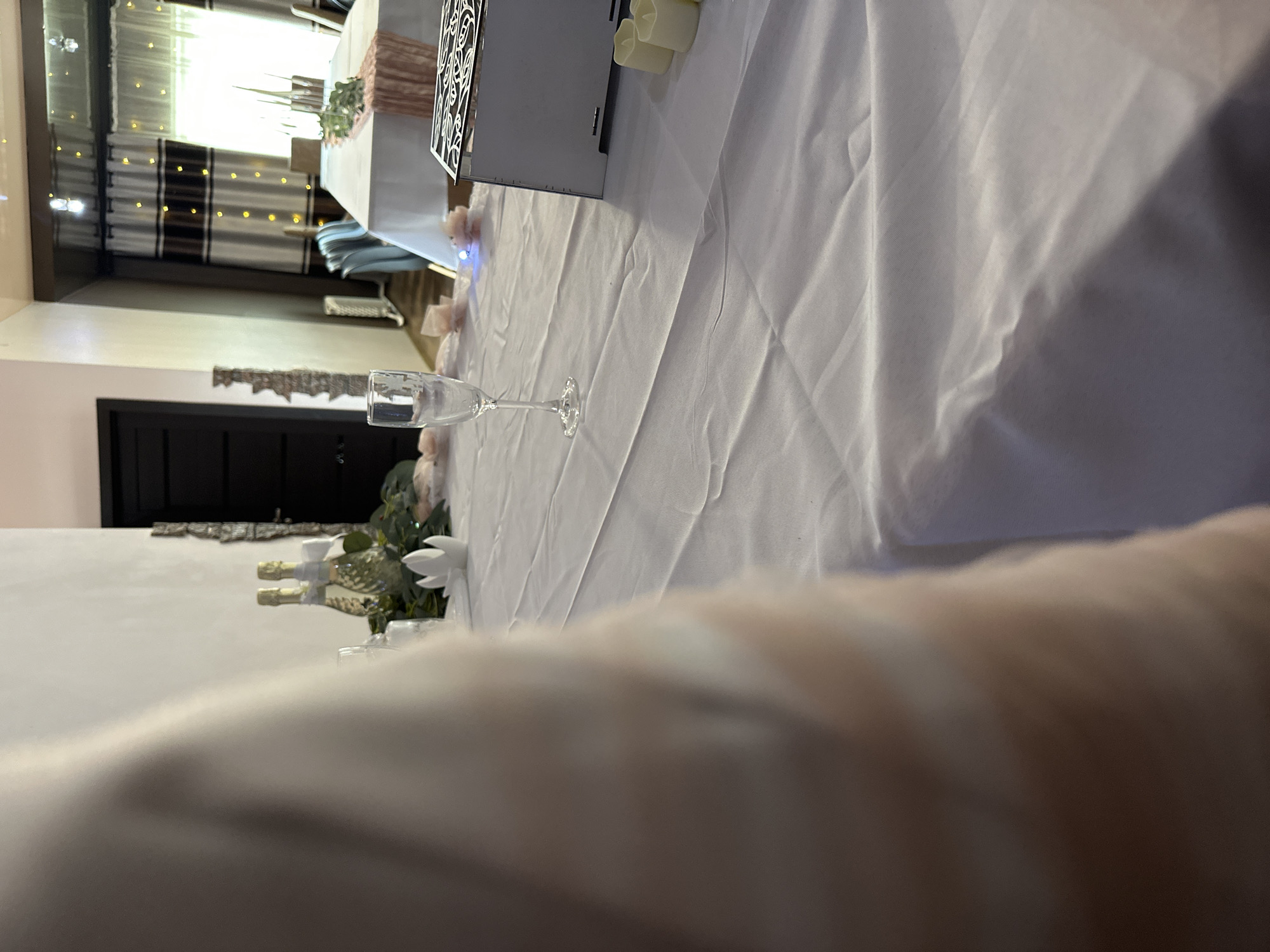 The image shows a wedding reception setting, likely indoors, with a long white tablecloth-covered table occupying a substantial portion of the frame. A champagne flute is prominently placed on the table, near several small champagne bottles and white floral decorations. Further along the table, a decorative box and faux candles can be seen. In the background, the venue is partially visible, featuring a dark door and decorative lighting and curtains. The location is given as the city of Unknown, Unknown. An arm partially obscures the bottom of the image.