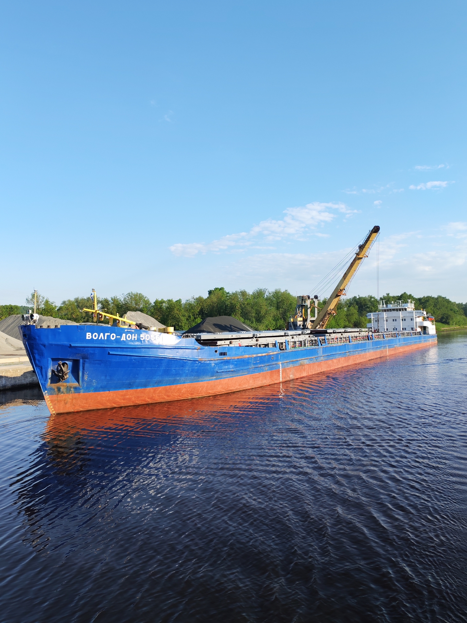 The image shows a blue and orange cargo ship docked near the riverbank in Yakhroma, Russia. The ship, named "VOLGO-DON 5055," is loaded with dark material, possibly coal or gravel. A yellow crane is mounted on the deck. The surrounding environment includes a concrete dock and a wooded area. The sky is blue with light clouds. The river water is dark with visible ripples.
