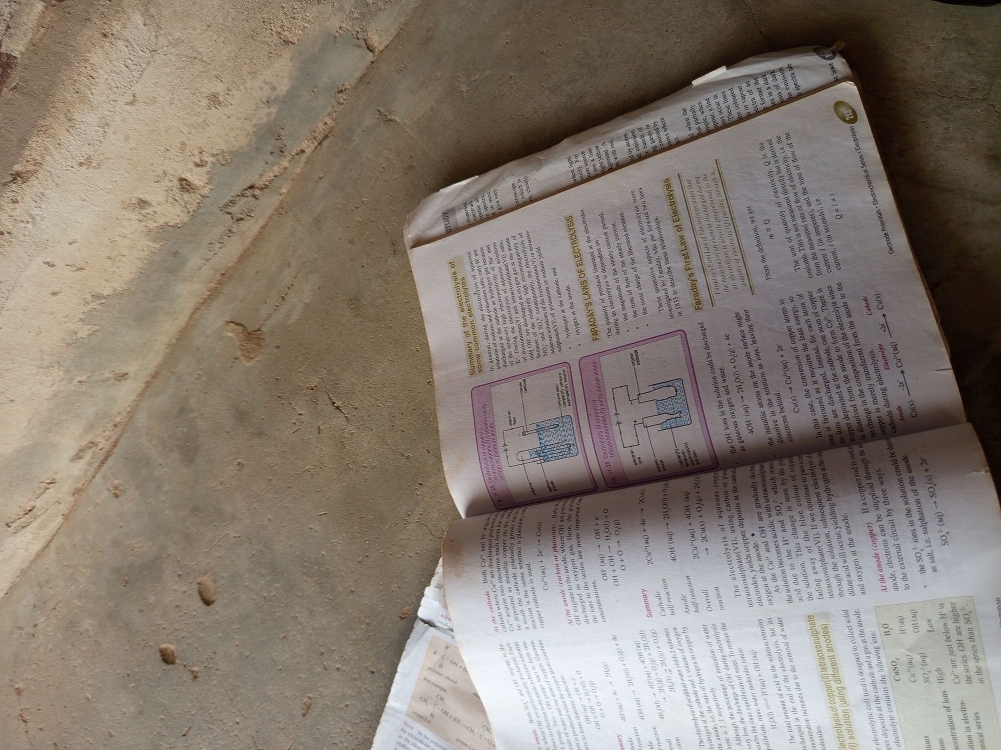 The image shows an open book with text and diagrams related to chemistry, specifically electrolysis. The pages are illuminated by natural light, casting shadows. The book is resting on a rough, concrete-like surface. One page details the "Summary of the electrolysis of some common electrolytes," including reactions and conditions. Another section discusses "FARADAY'S LAWS OF ELECTROLYSIS," with diagrams illustrating the process. The text is primarily in English. The specific location in Jalingo, Nigeria cannot be determined from the provided image.
