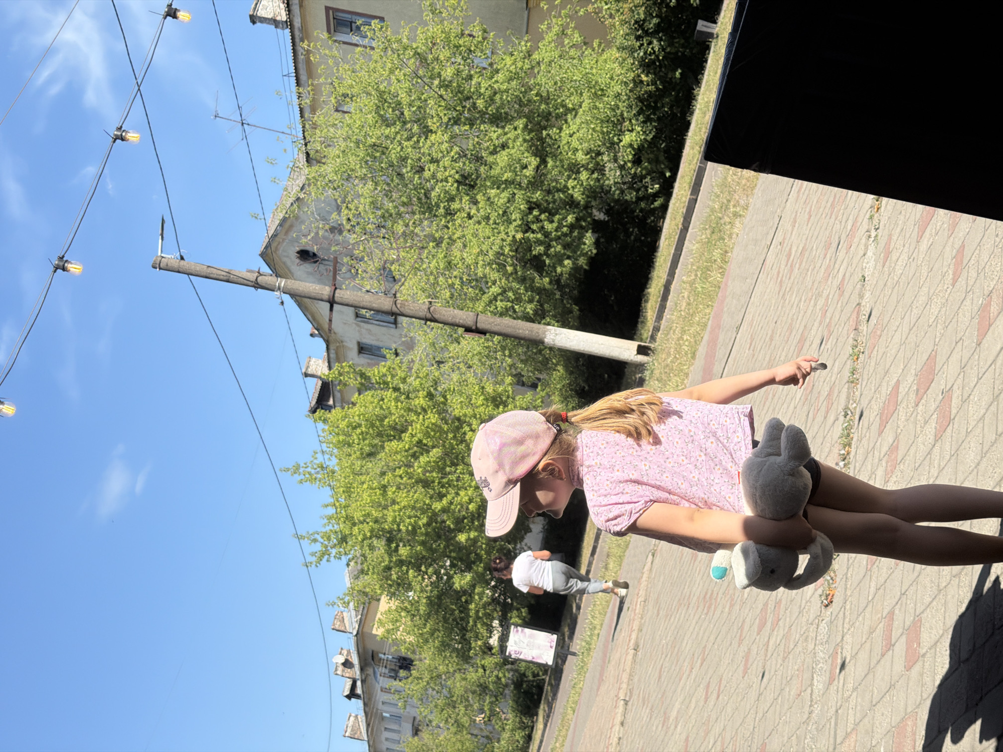 In an outdoor urban setting, a young girl, viewed from behind and the side, walks on a paved pathway. She wears a pink patterned short-sleeved shirt and dark shorts, with a pink baseball cap on her head. She carries a light gray plush toy animal in her right arm. Her blonde hair is styled in a ponytail. Further up the pathway, another person, dressed in a white t-shirt and gray pants, is seen walking away from the camera. The environment includes a sidewalk with brick paving, a grassy area, trees, and buildings in the background. Overhead, a string of lights with visible bulbs hangs across the sky, and a utility pole stands prominently. The scene is illuminated by daylight, casting shadows on the pathway.