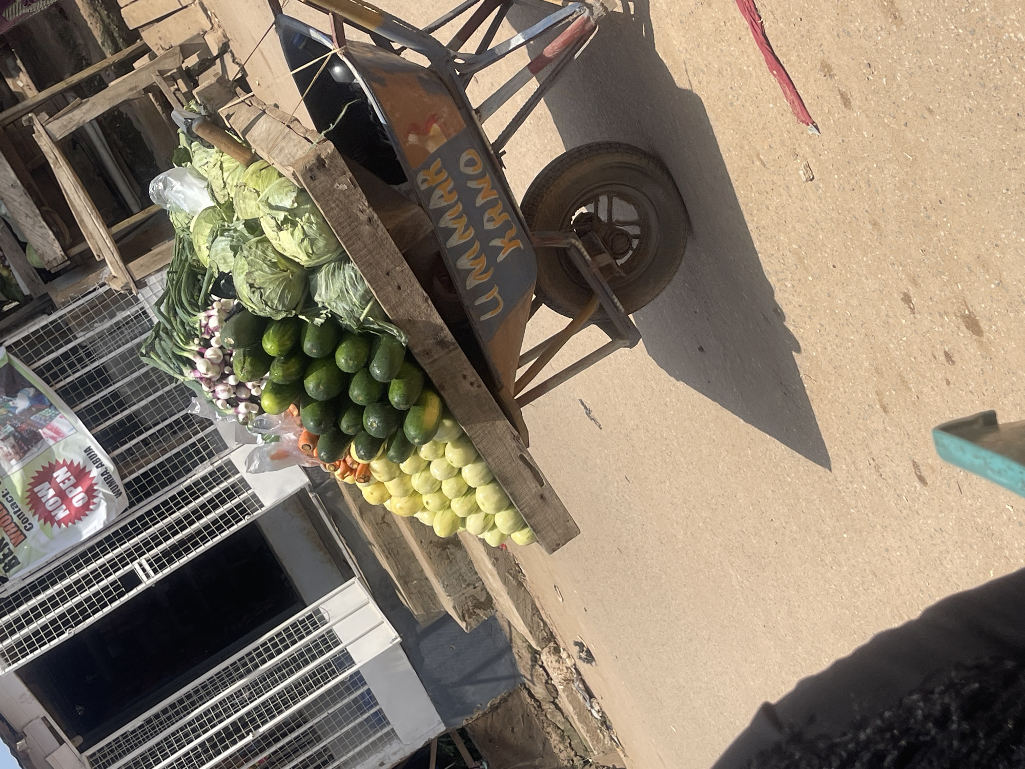 An outdoor market scene in Abuja, Nigeria, features a wooden cart filled with fresh produce, including green leafy vegetables like cabbage and lettuce, along with piles of avocados and yellow fruits. The cart is also laden with onions and carrots. The cart itself is a wheelbarrow with a metal basin painted in orange and dark grey hues, bearing the inscription "UMMAR KANO" in orange lettering. The background reveals a building with a white facade and metal grates on its windows and doors. A banner above the entrance reads "NOW OPEN" in bold red lettering with white outlines, and "WUMBA, ABUJA" is visible below. The ground is a paved road, illuminated by sunlight, casting shadows from the cart and other unseen objects. The image has a slight tilt, as if captured from a moving perspective.